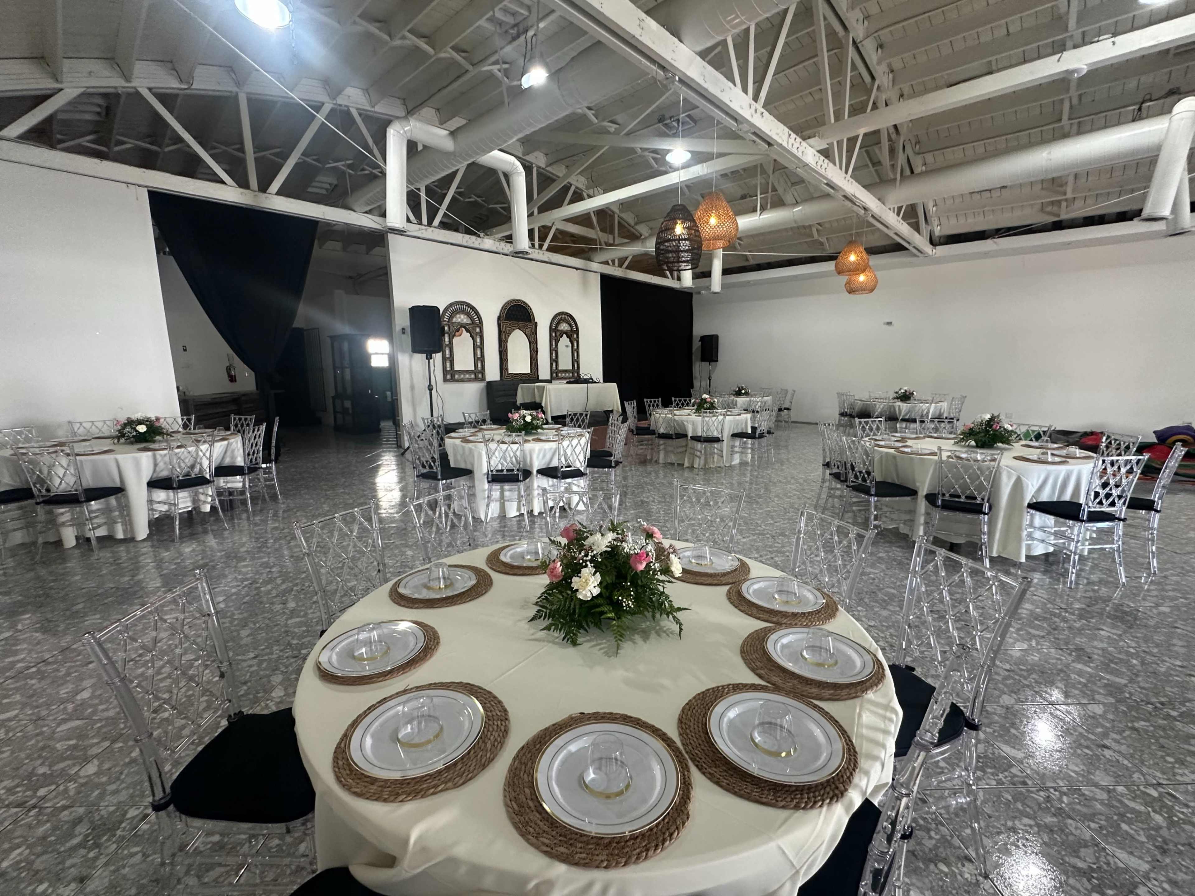 The image shows a spacious event venue with circular tables neatly set with dinnerware and floral centerpieces, along with a stage area and light fixtures hanging from the ceiling.