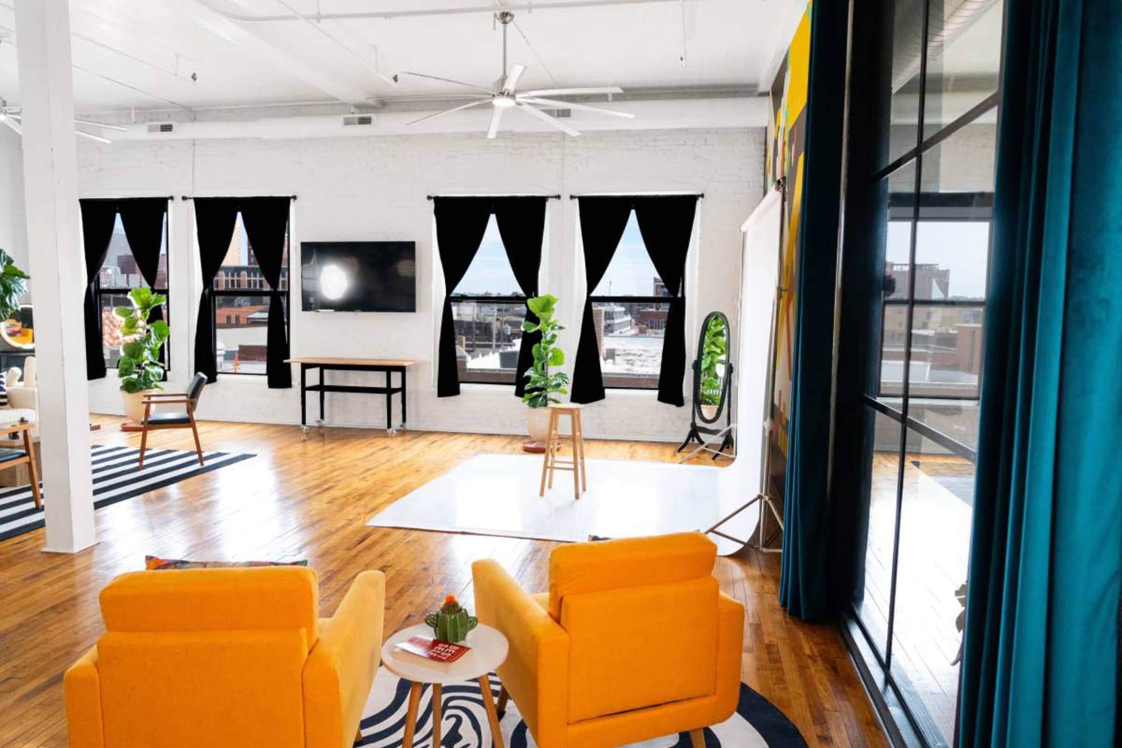 The image shows a bright, spacious interior with two orange chairs facing each other, large windows draped with black curtains, and wooden flooring.