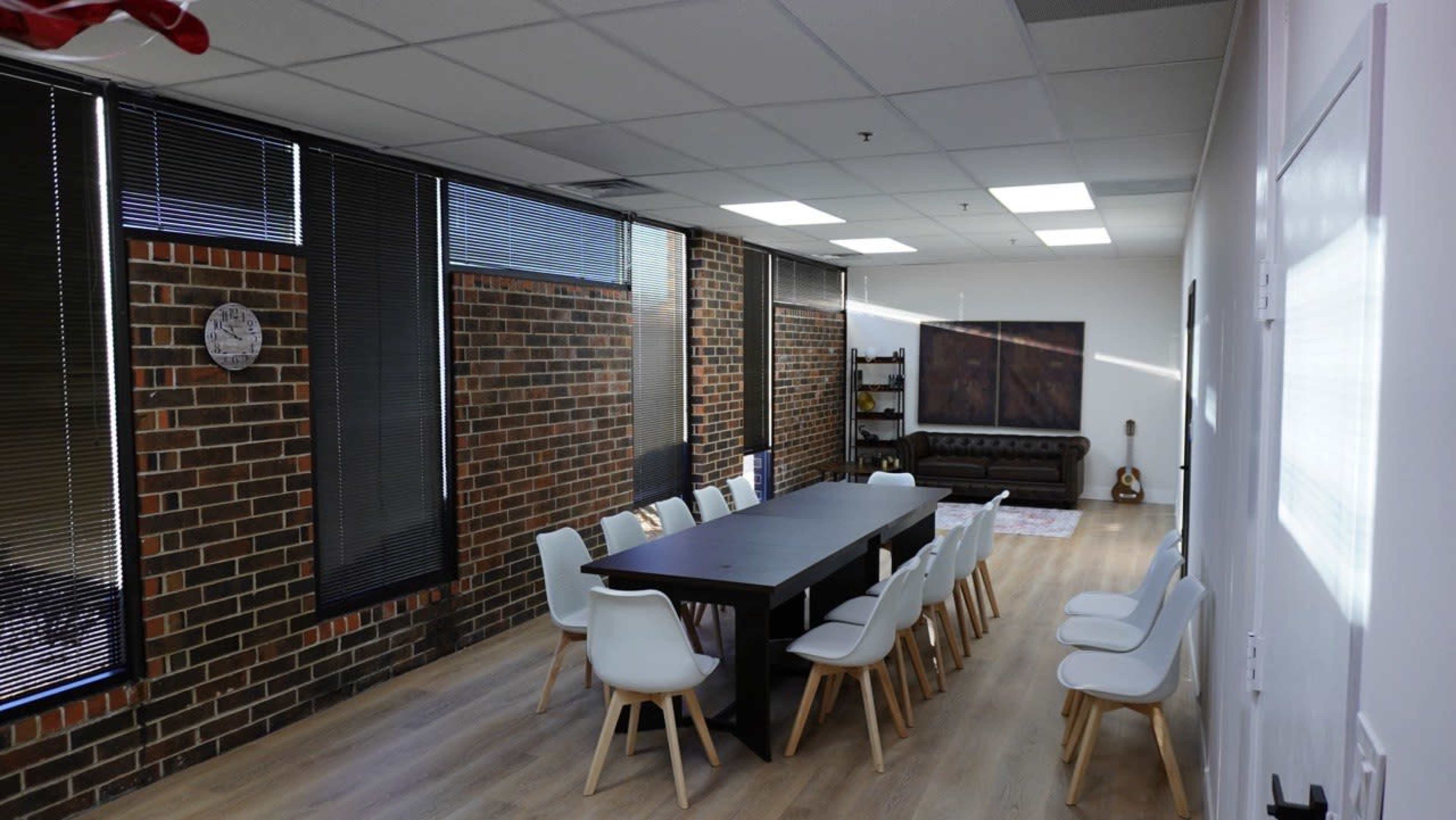 The image shows a modern meeting room featuring a long black table surrounded by white chairs, with large windows and brick walls.