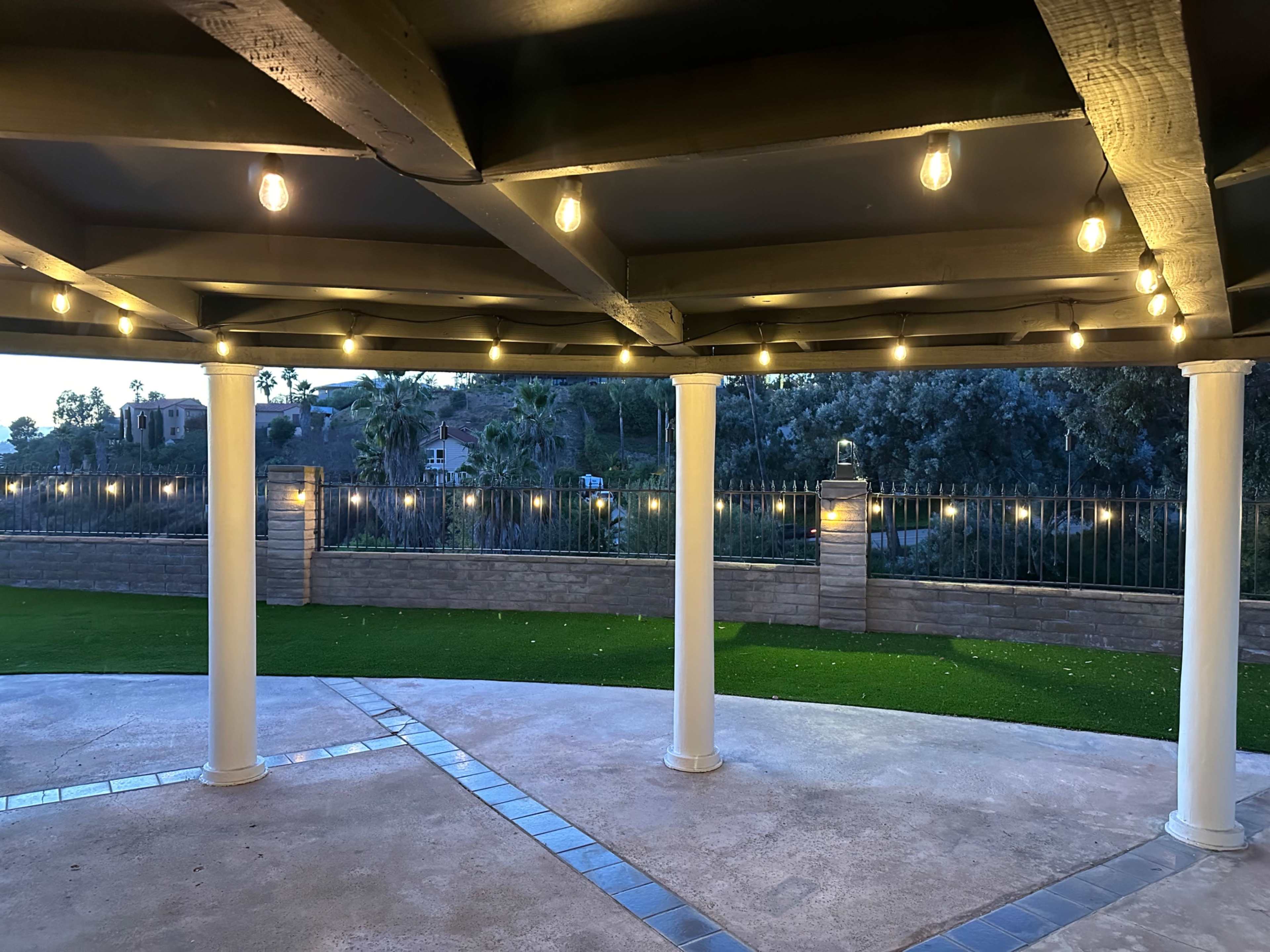 A patio area with a ceiling featuring string lights and white columns, overlooking a landscaped yard with a fence in the background.