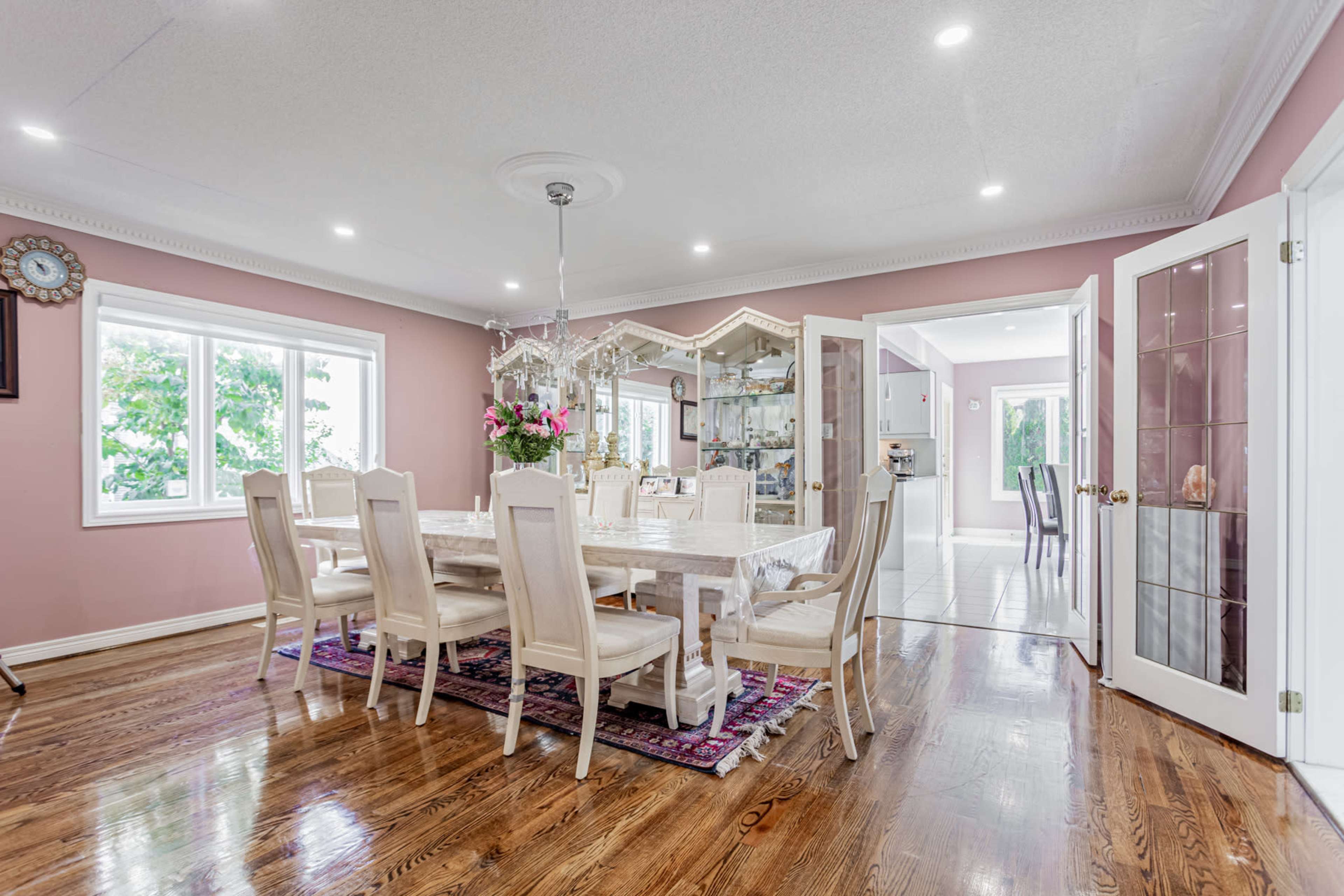 A spacious dining room features a large table surrounded by upholstered chairs, with a chandelier overhead and windows letting in natural light.