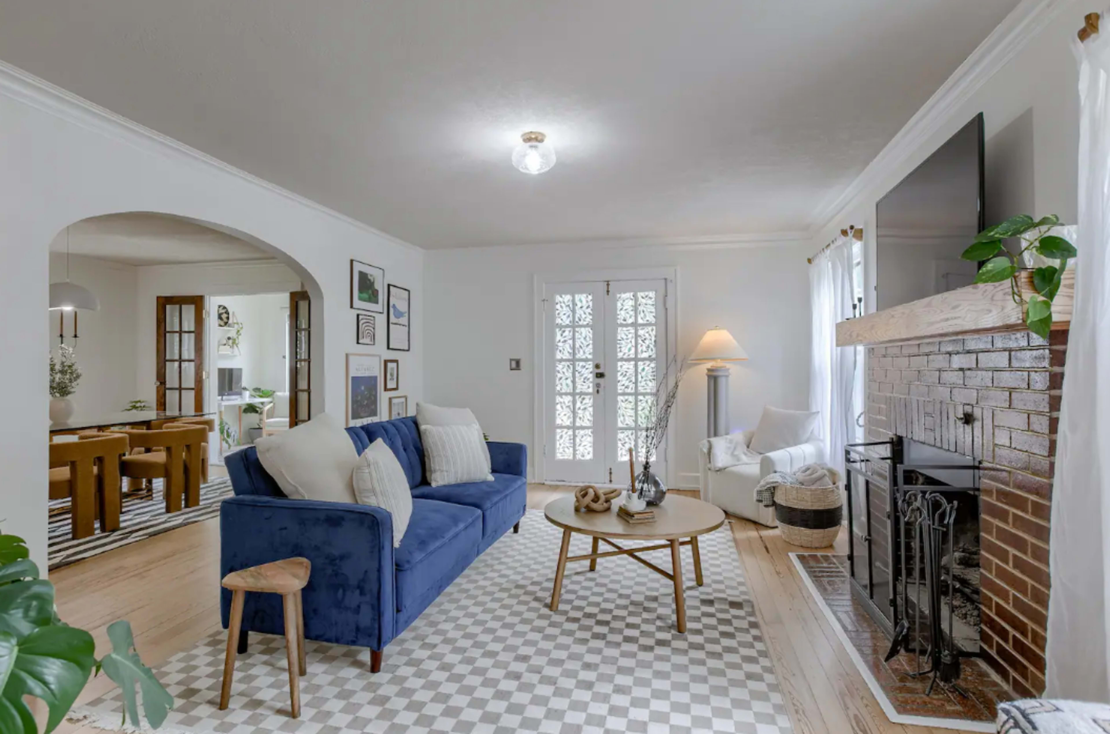 A cozy living room features a blue sofa, a round coffee table, and a fireplace, with light streaming through large windows and an adjoining dining area visible in the background.