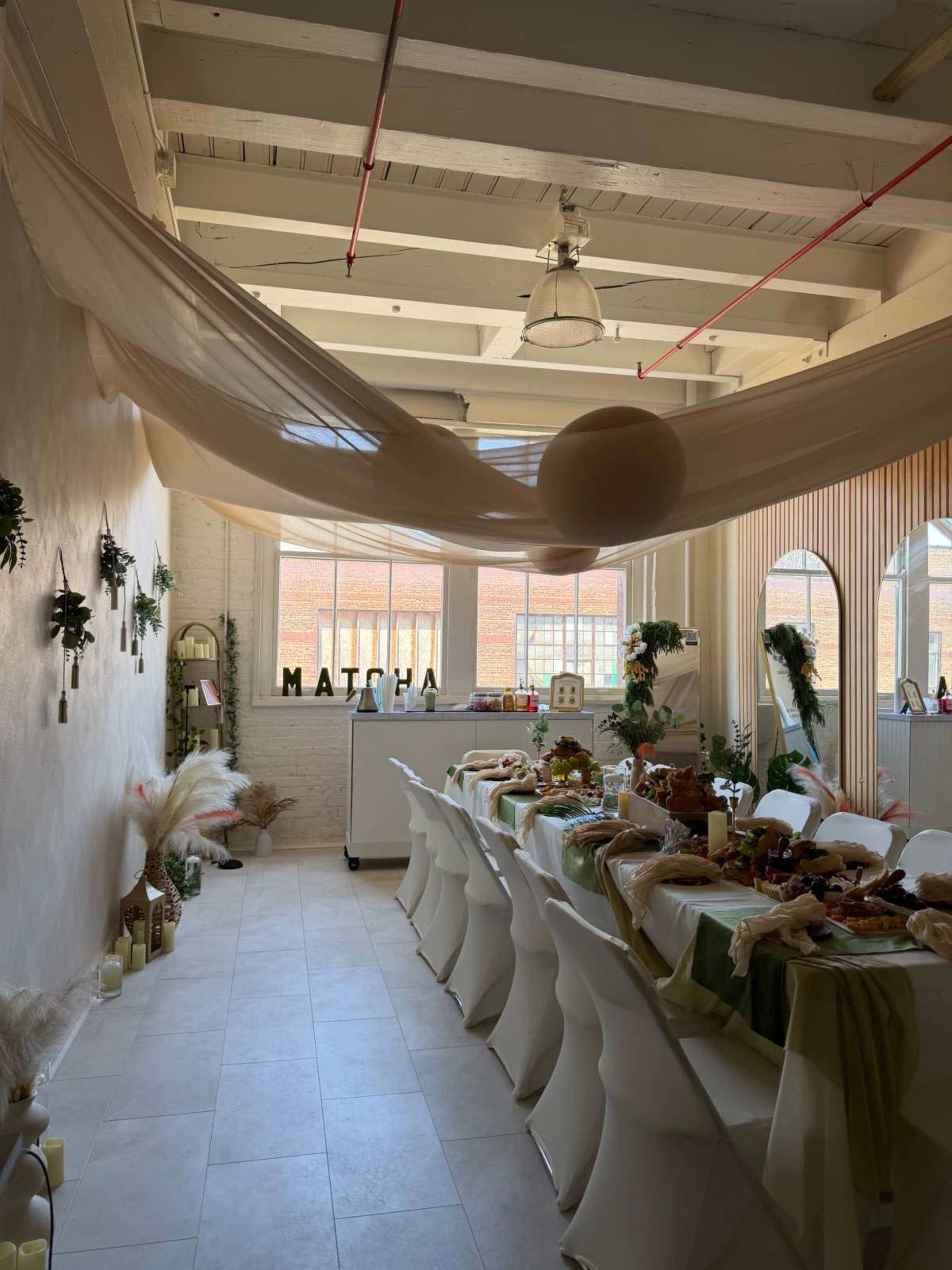 The image shows a decorated dining area with a long table set for an event, adorned with various floral arrangements and draped fabric overhead.