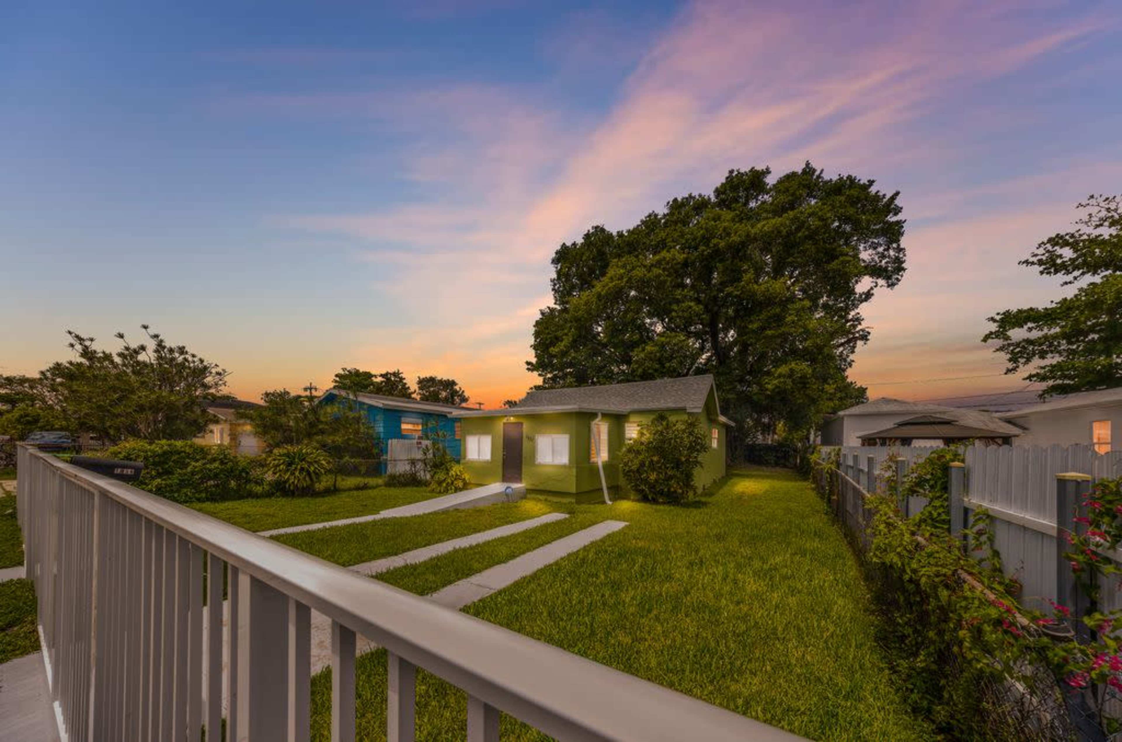 A house with a green lawn and trees is viewed from a sidewalk at sunset, with a colorful sky in the background.