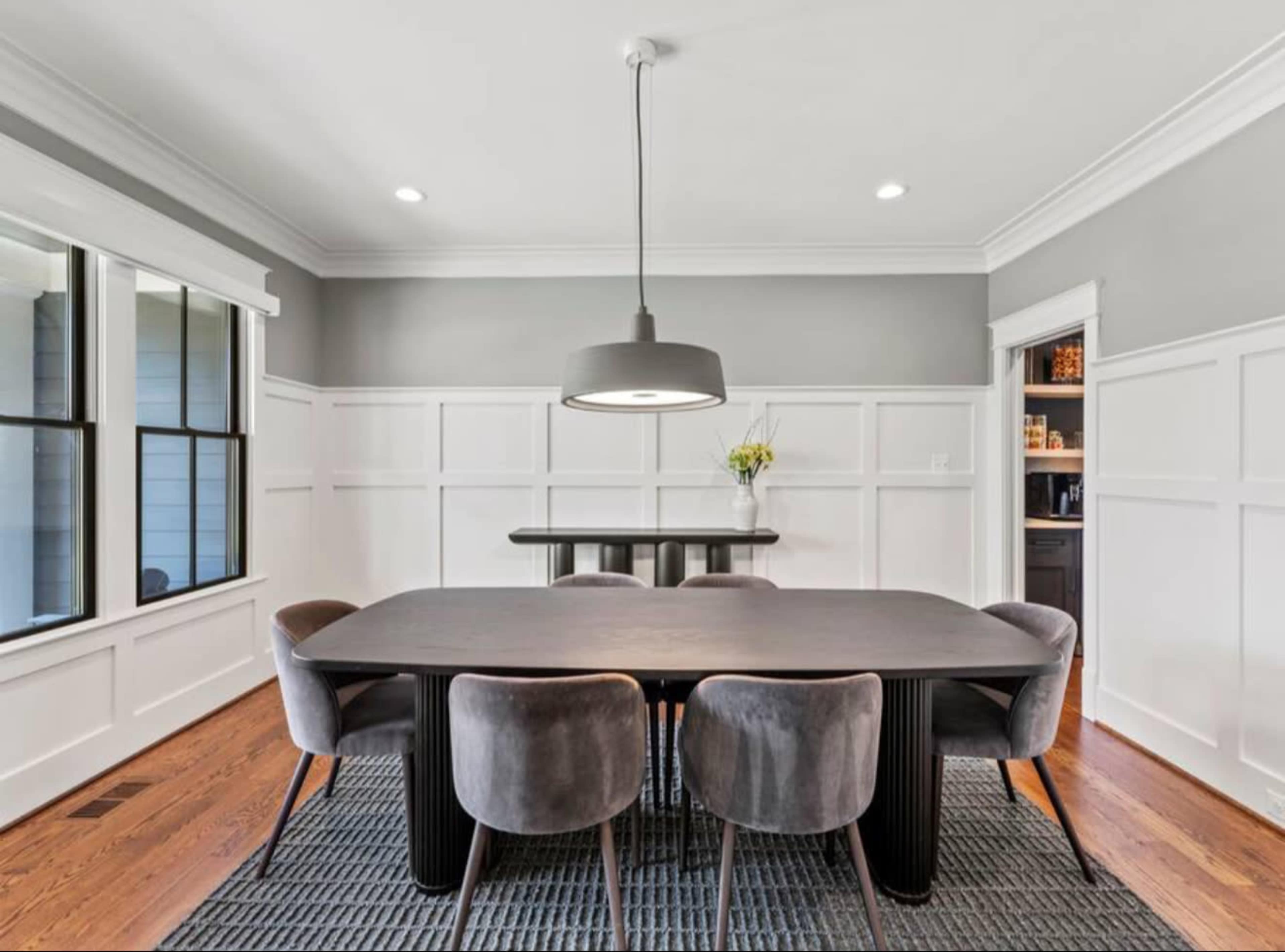 A modern dining room features a long black table surrounded by gray upholstered chairs, with a pendant light hanging above and a built-in cabinet visible in the background.