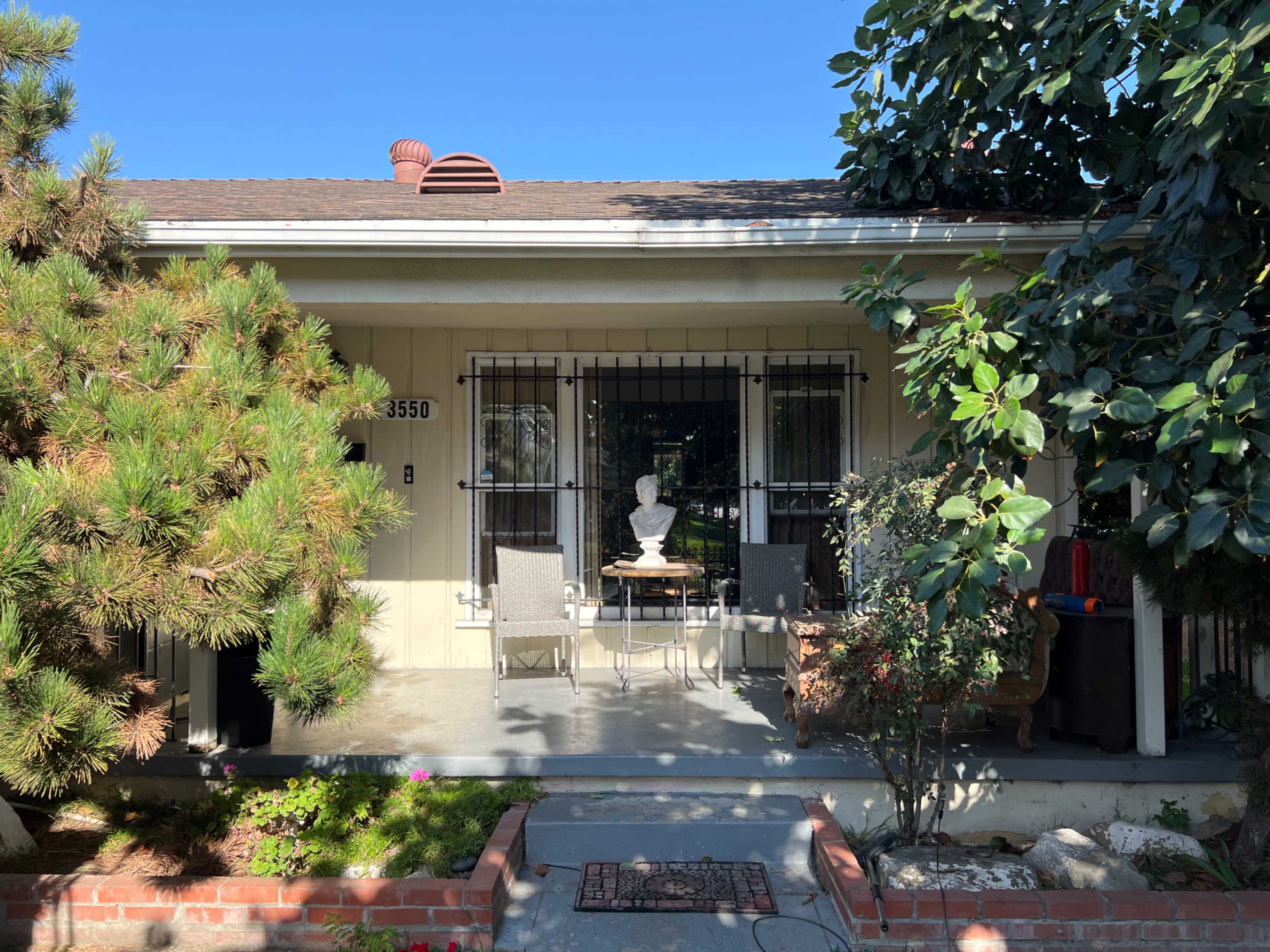 The image shows a front porch with two chairs and a small table, bordered by a landscaped area with shrubs and a statue.