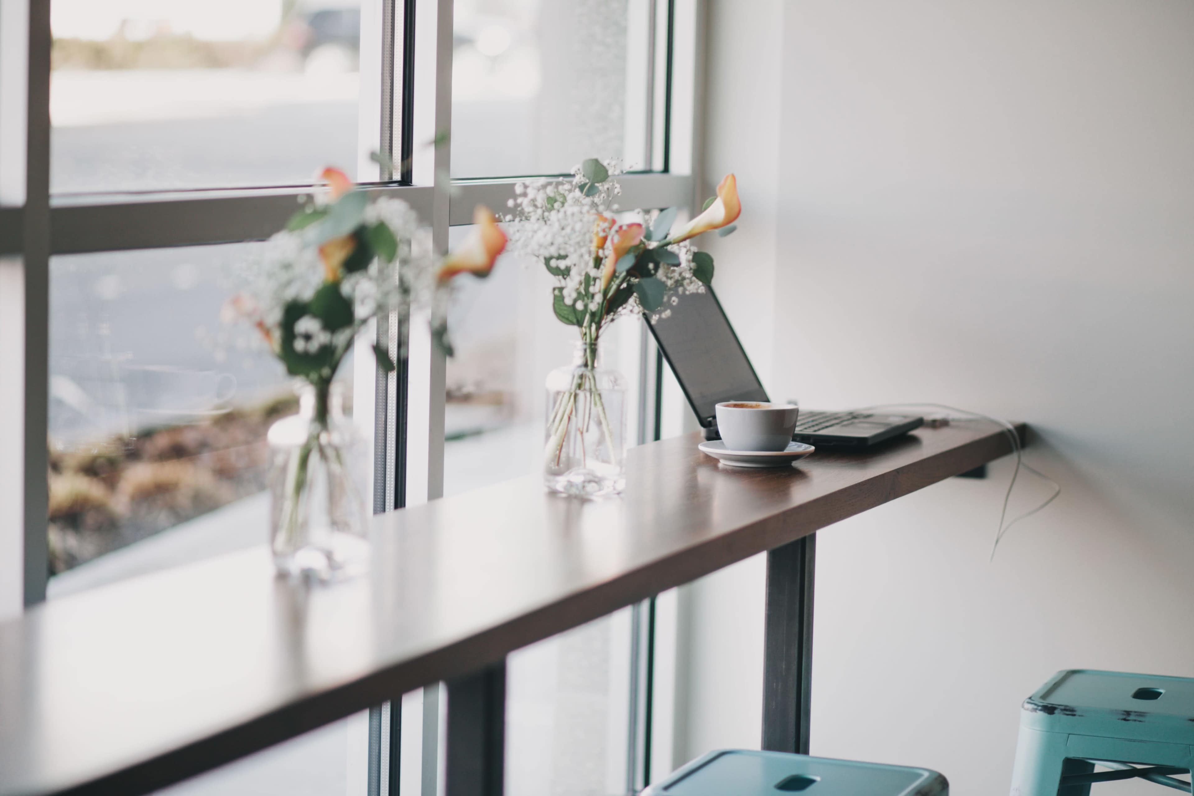A narrow wooden table beside large windows features two vases with flowers, a laptop, and a cup of coffee.