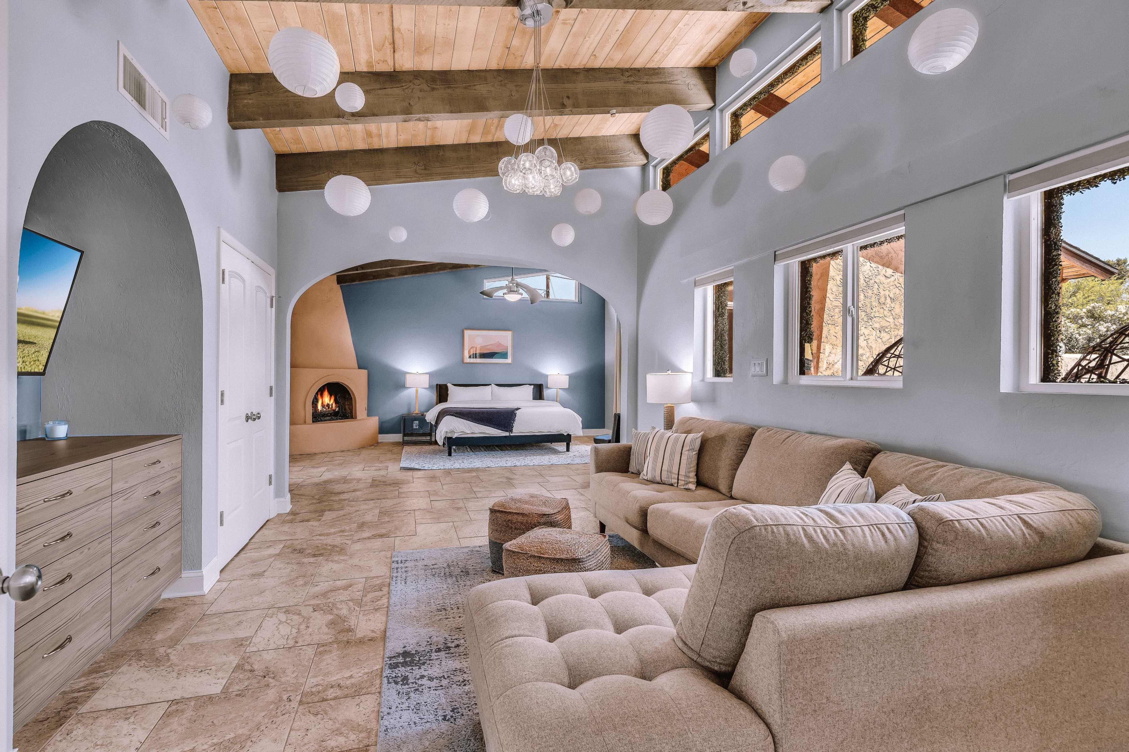 A spacious living area with a beige sectional sofa, a wooden dresser, and large windows that illuminate a bedroom in the background featuring a bed and a fireplace.