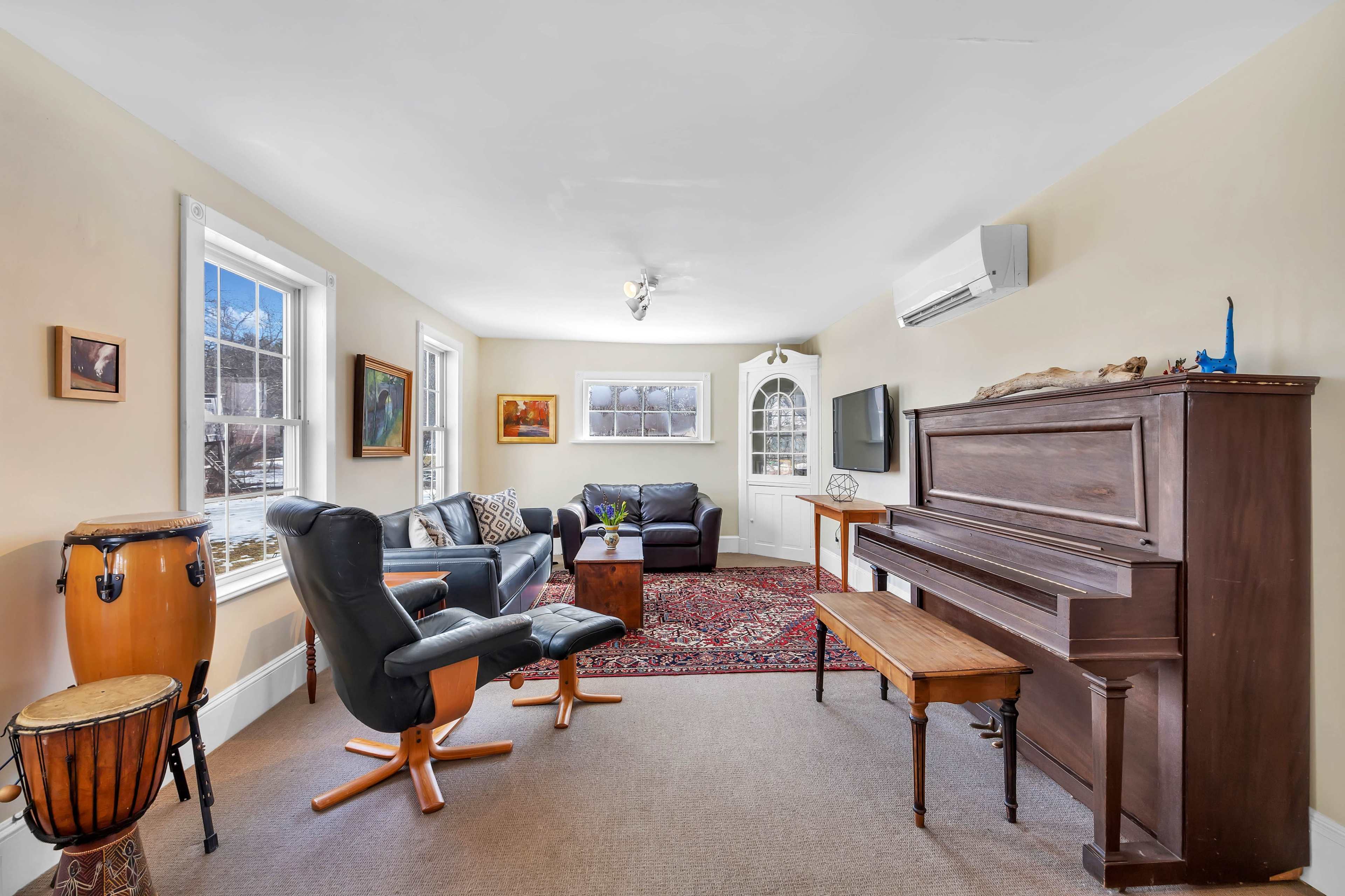 The image shows a living room with a piano, seating arrangements, and windows that provide natural light.