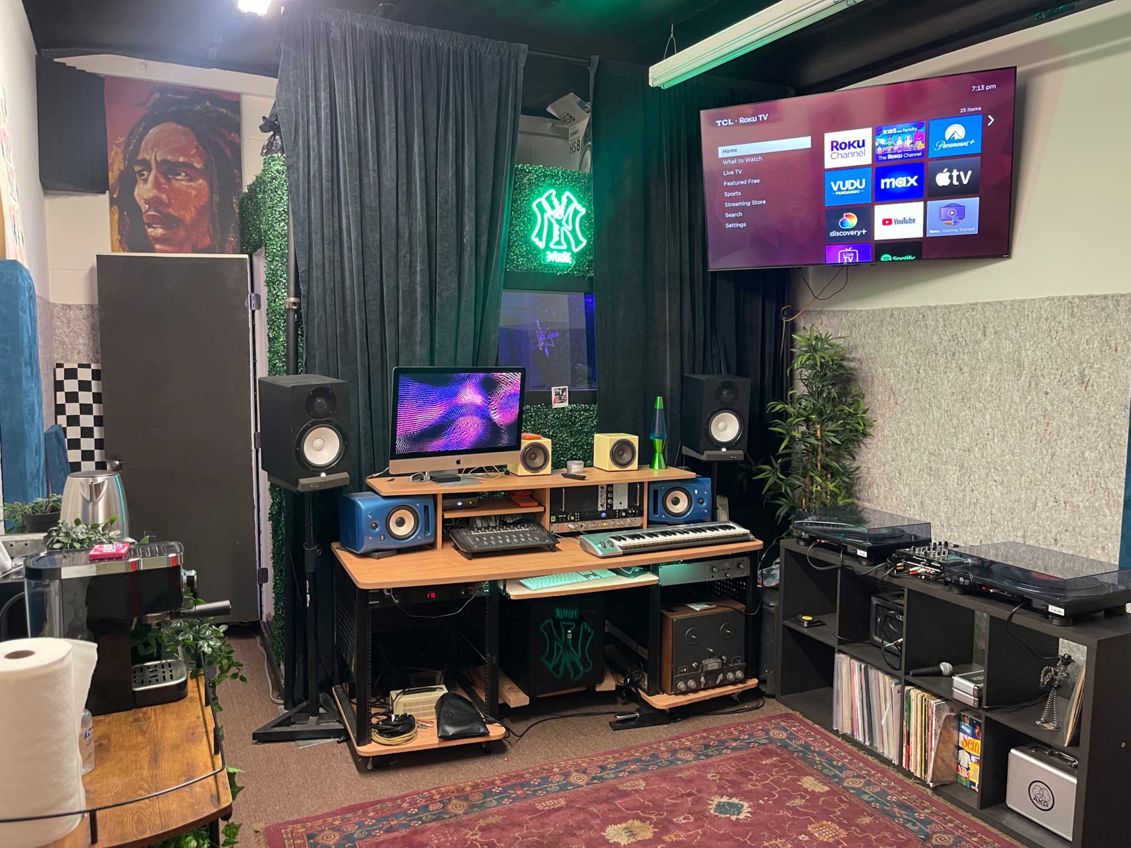 A music studio with a computer desk featuring audio equipment, speakers, and a vinyl record player, surrounded by greenery and colorful wall art.