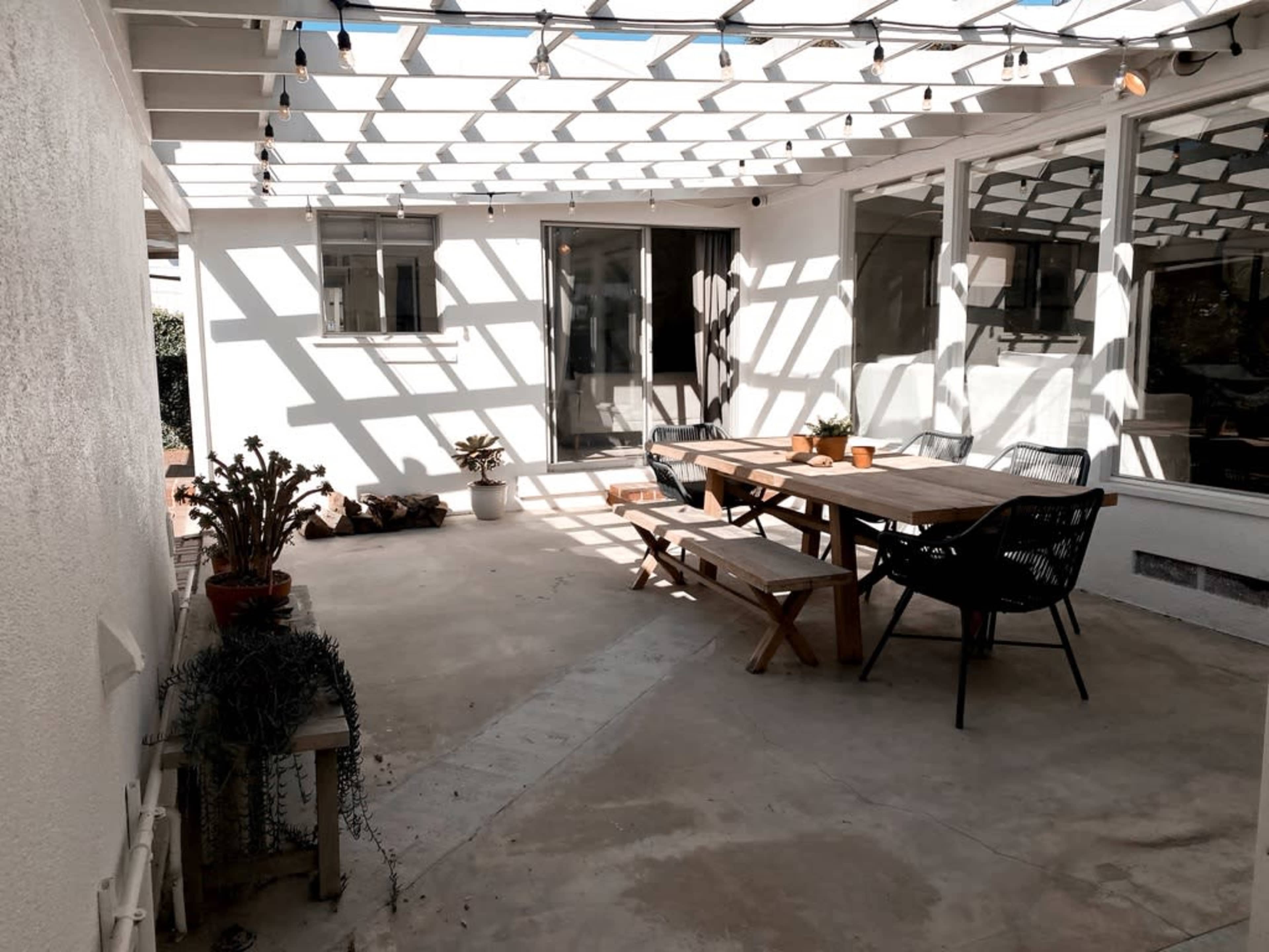 A covered patio area with a wooden table and chairs, surrounded by potted plants and sunlight casting geometric shadows on the floor.