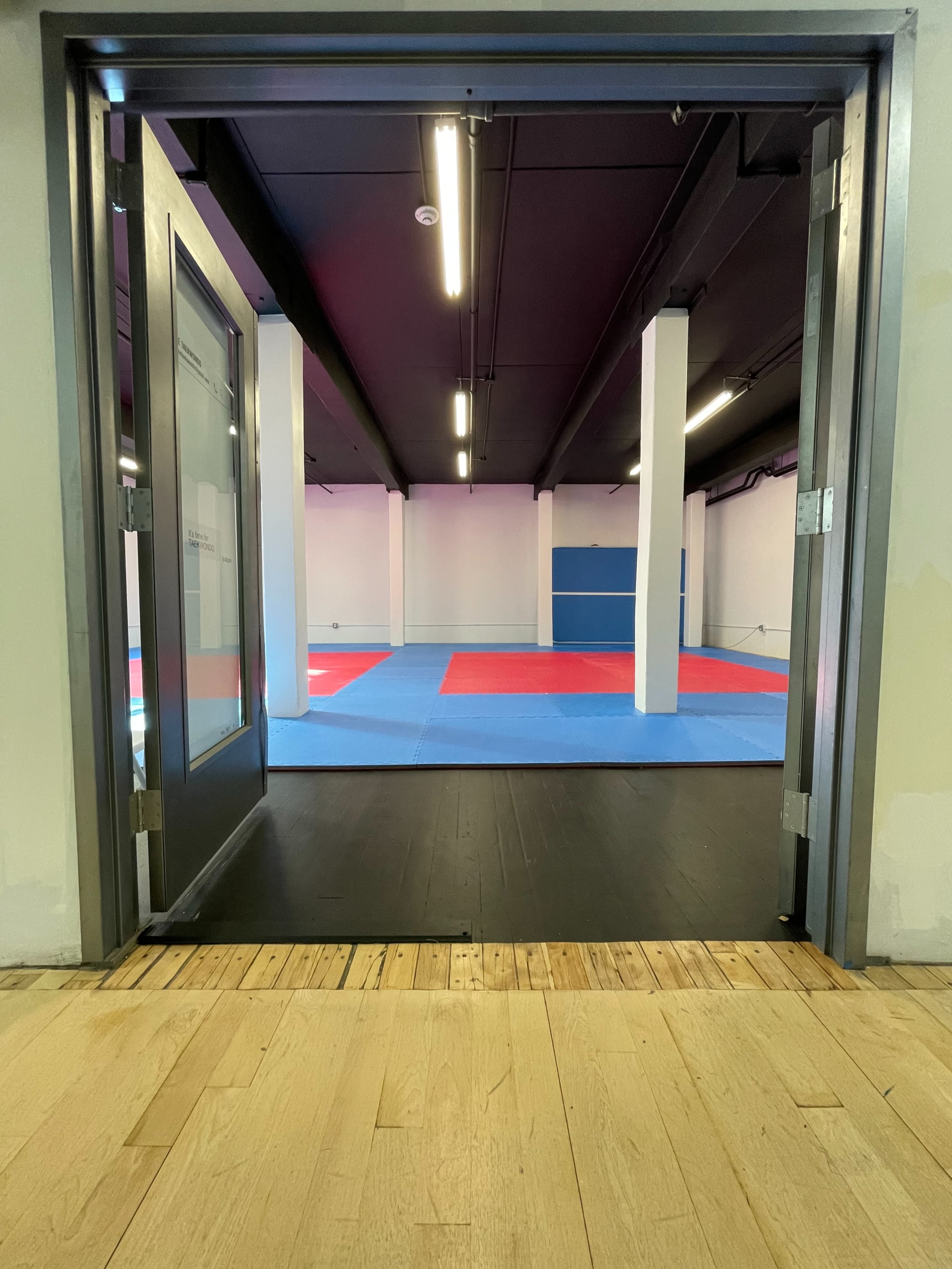 An open doorway leading into a large martial arts training room with blue and red mats.