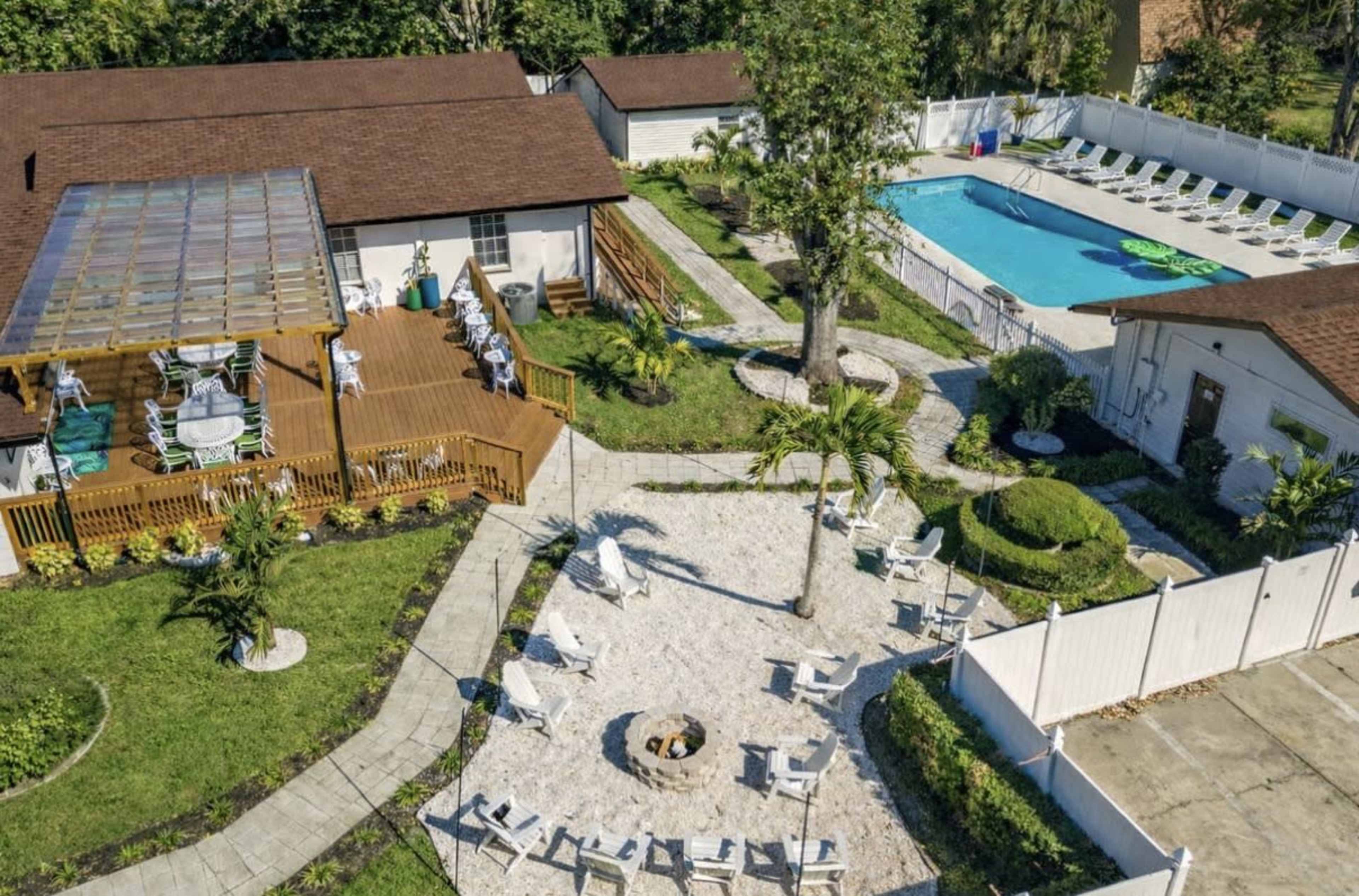 The image shows an aerial view of a property featuring a swimming pool, a wooden deck area, lounge chairs, and a landscaped garden with a fire pit.