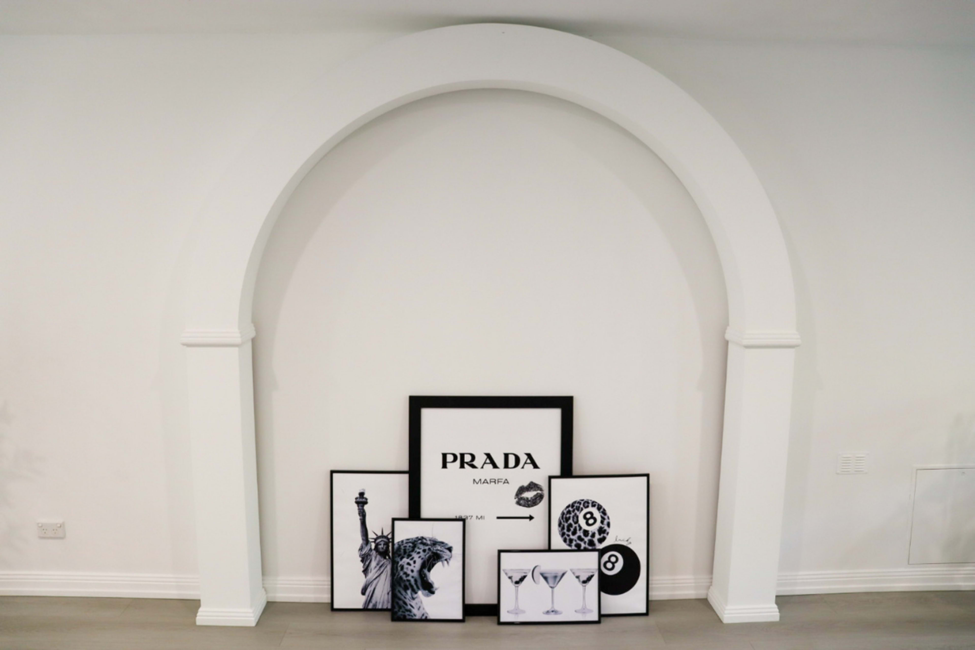 The image shows an arched alcove on a wall displaying a collection of framed black and white artworks, including a Prada poster and various graphic prints.