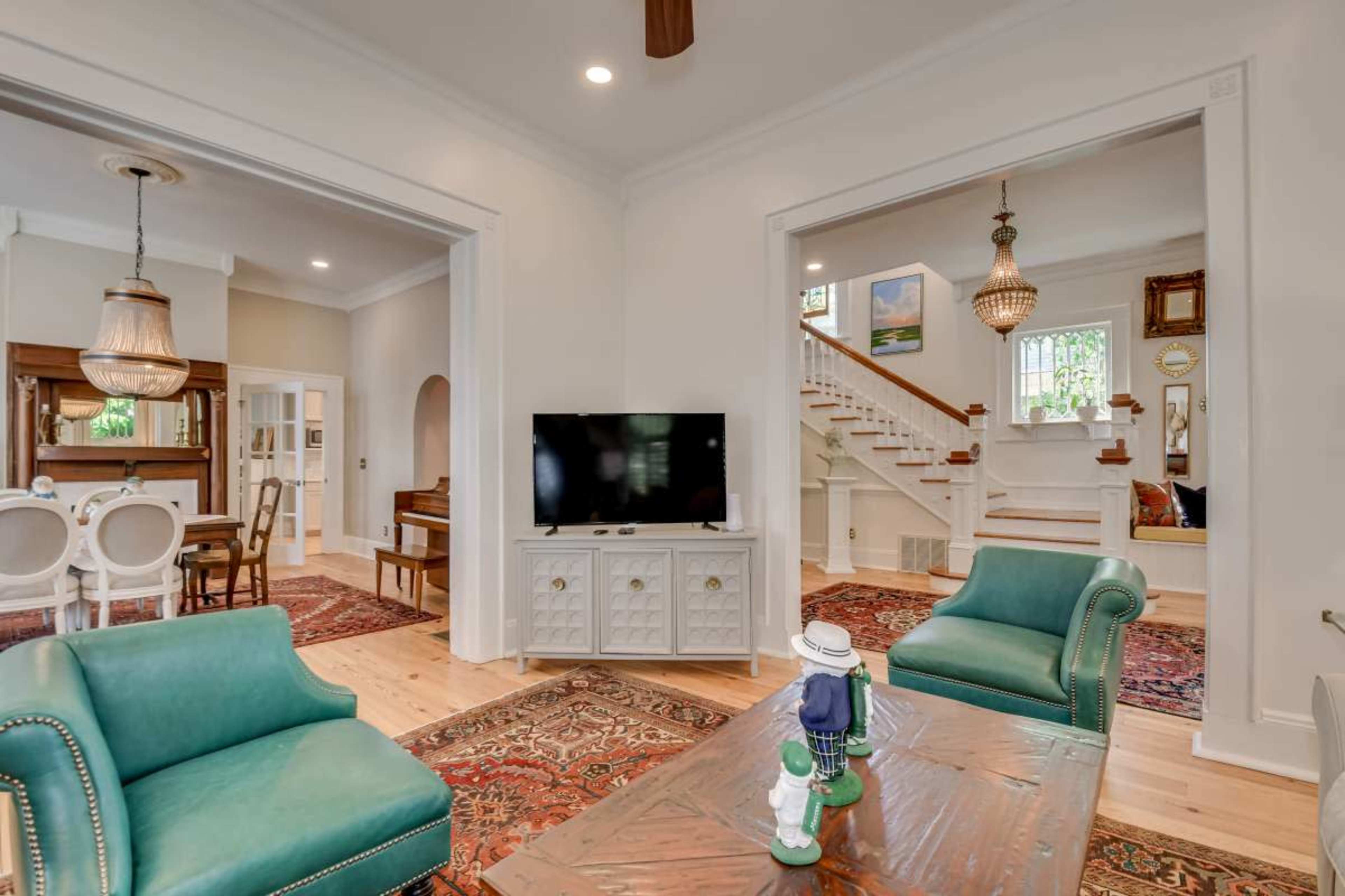 The image shows a living room with two green chairs and a wooden coffee table, featuring a television and an entrance to a staircase and another room.