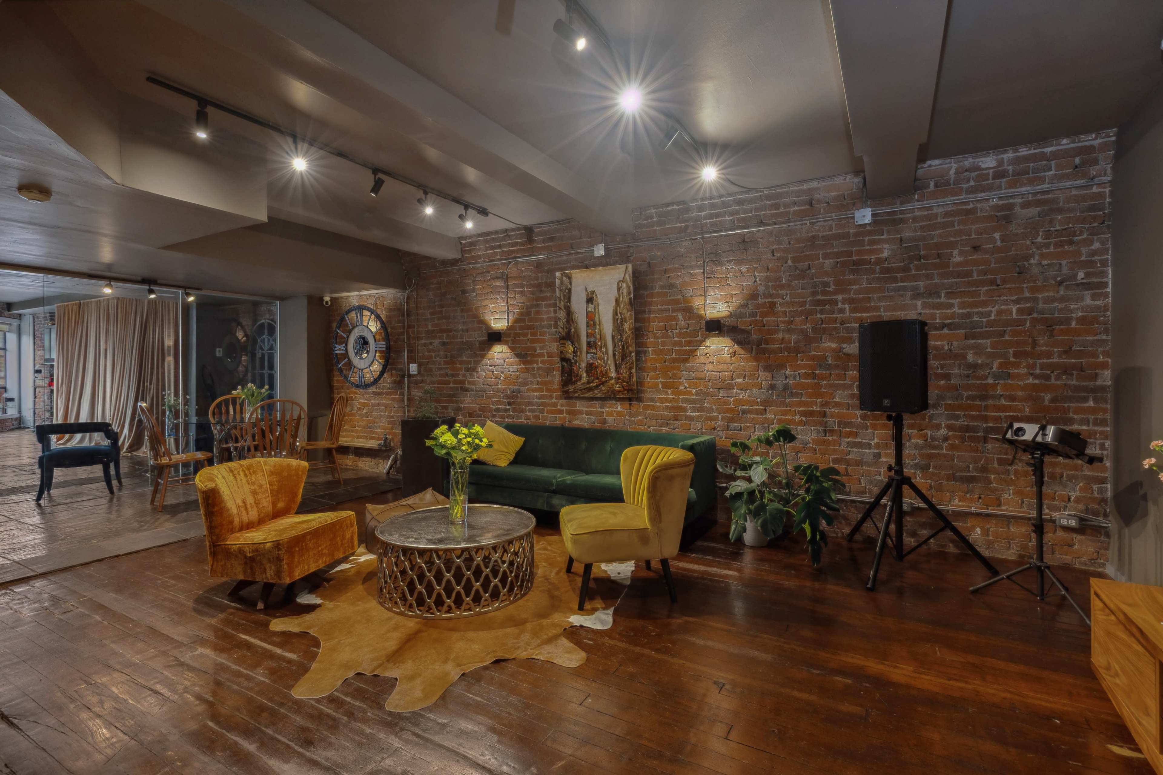 The image shows a rustic interior space featuring exposed brick walls, a green sofa, two colorful chairs, a round coffee table on a cowhide rug, and a small audio setup.