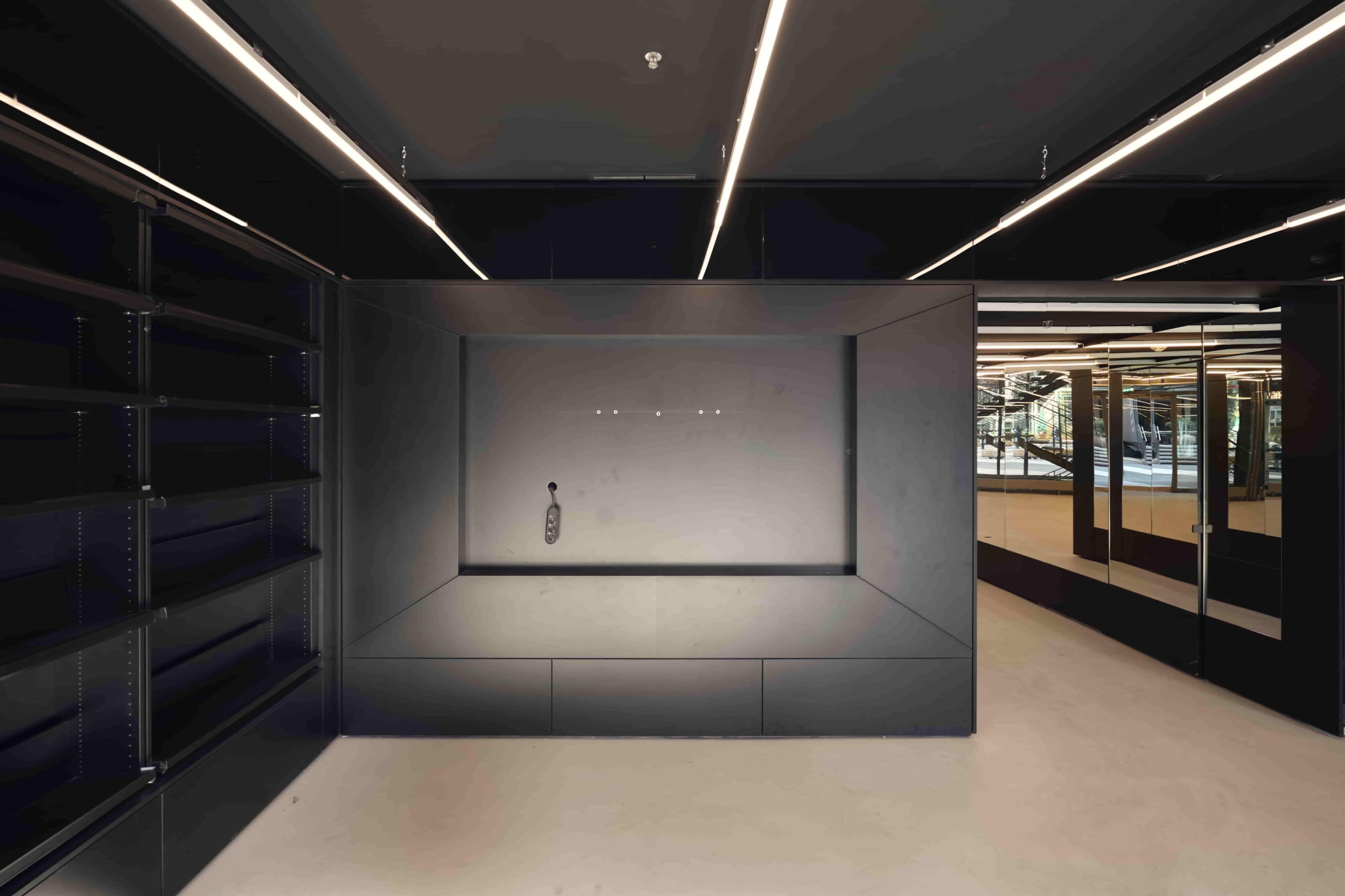The image shows an empty, modern retail space featuring black shelving units along the walls and a central display area with sleek lighting.