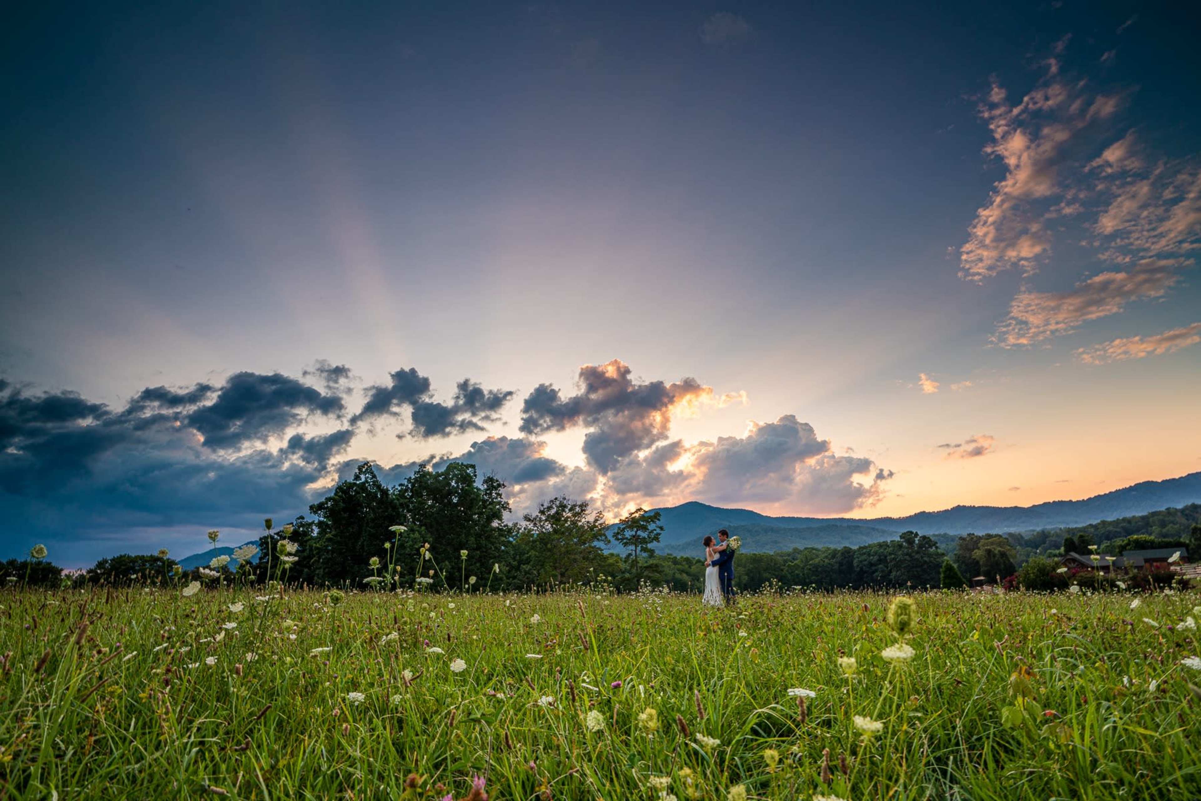 A couple embraces in a grassy field under a sky filled with clouds and rays of sunlight at sunset.