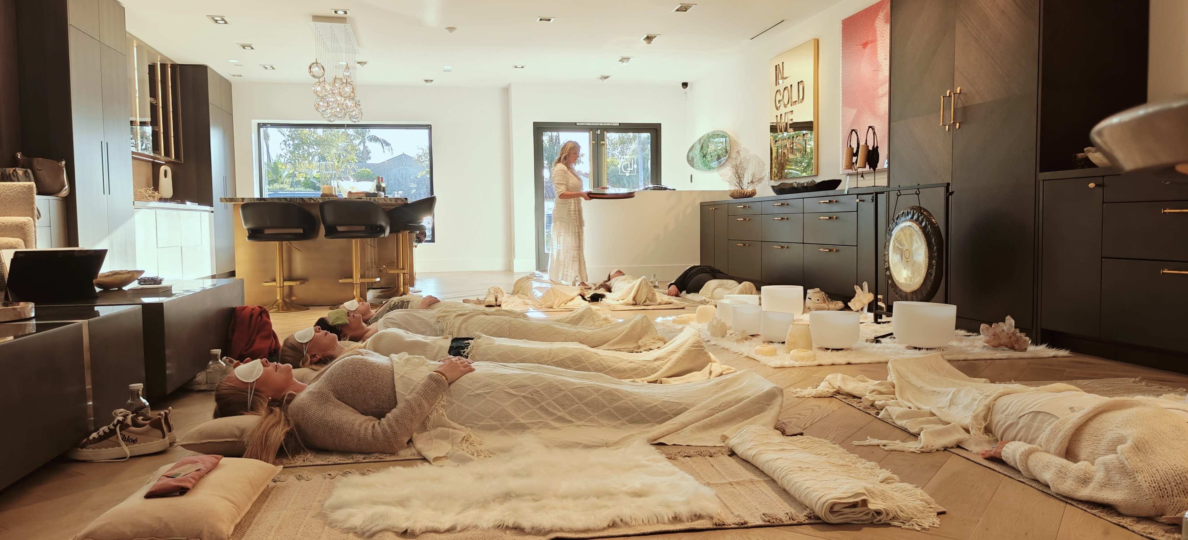 A group of people lies on mats on the floor of a serene room, while a woman stands behind a counter, and various objects are arranged around the space.