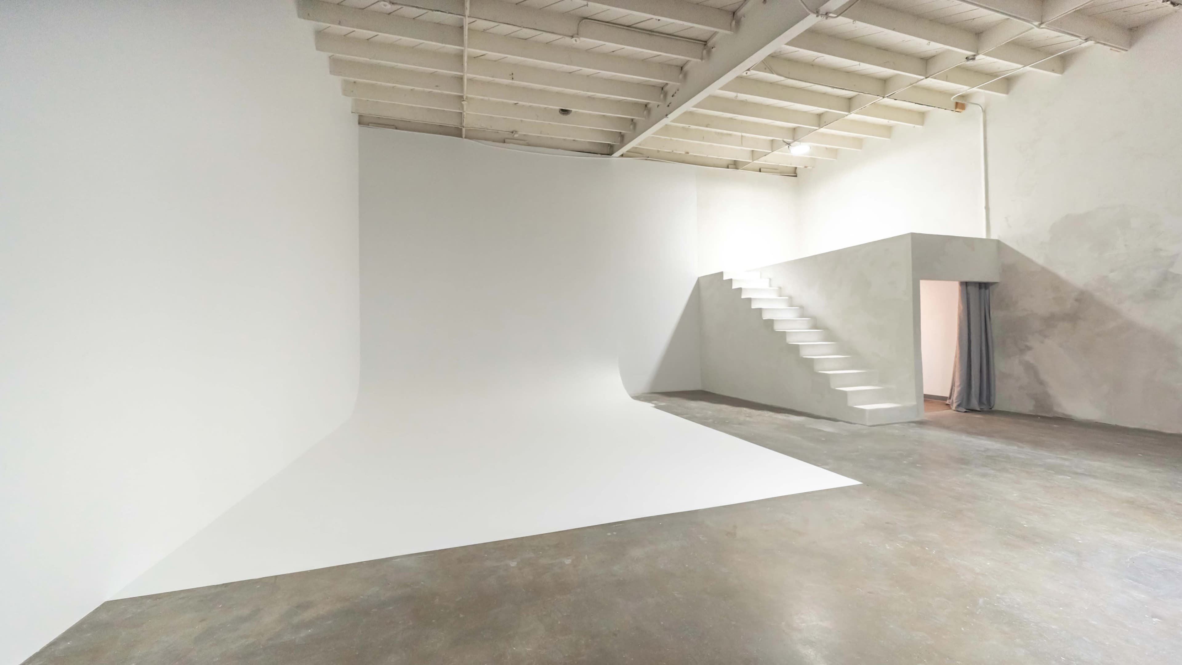 The image depicts a spacious, empty room with white walls, a white curved backdrop, a set of stairs, and a doorway.