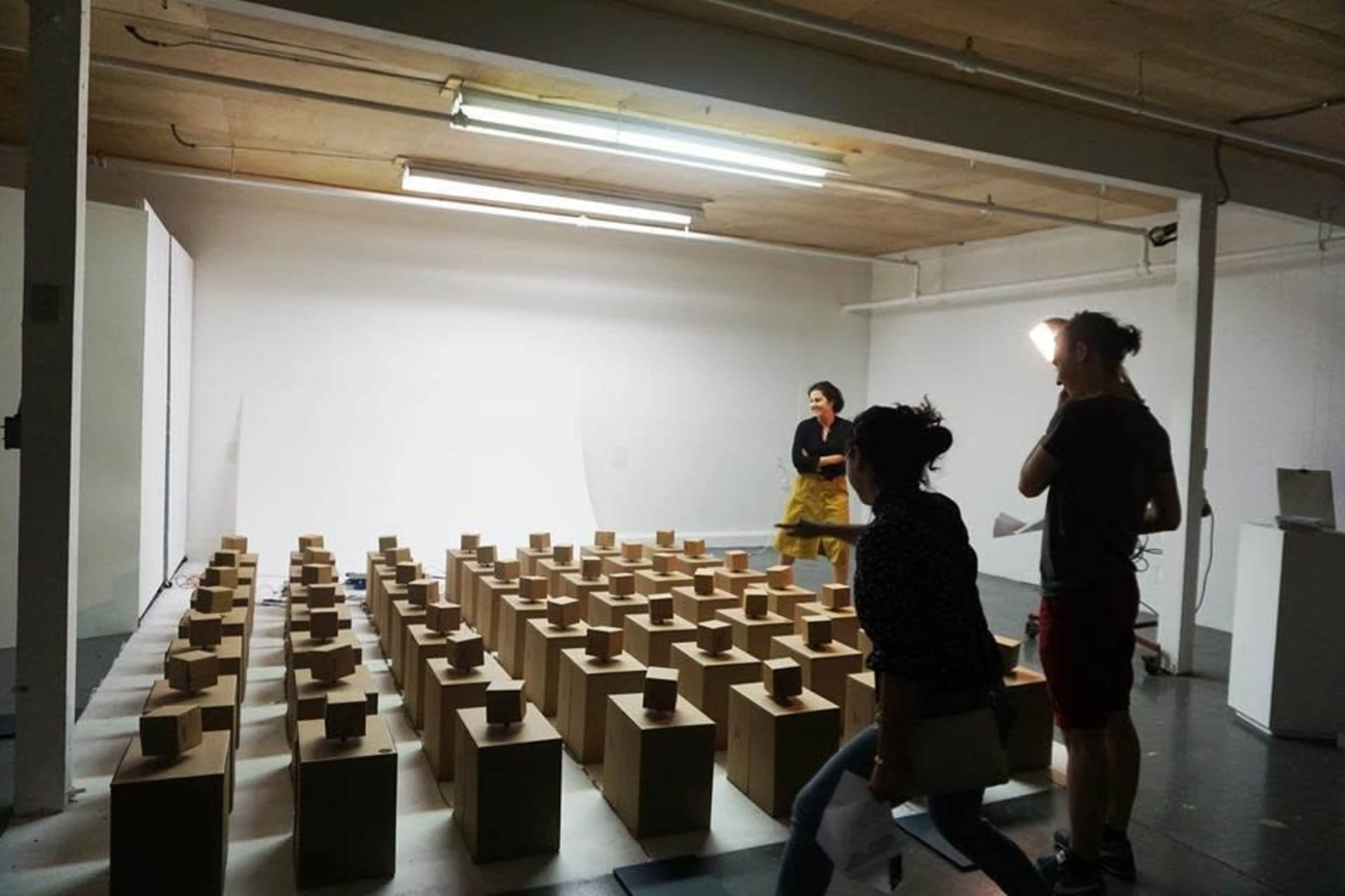 A studio space is organized with numerous wooden boxes arranged in rows, while two people are positioned in the background.