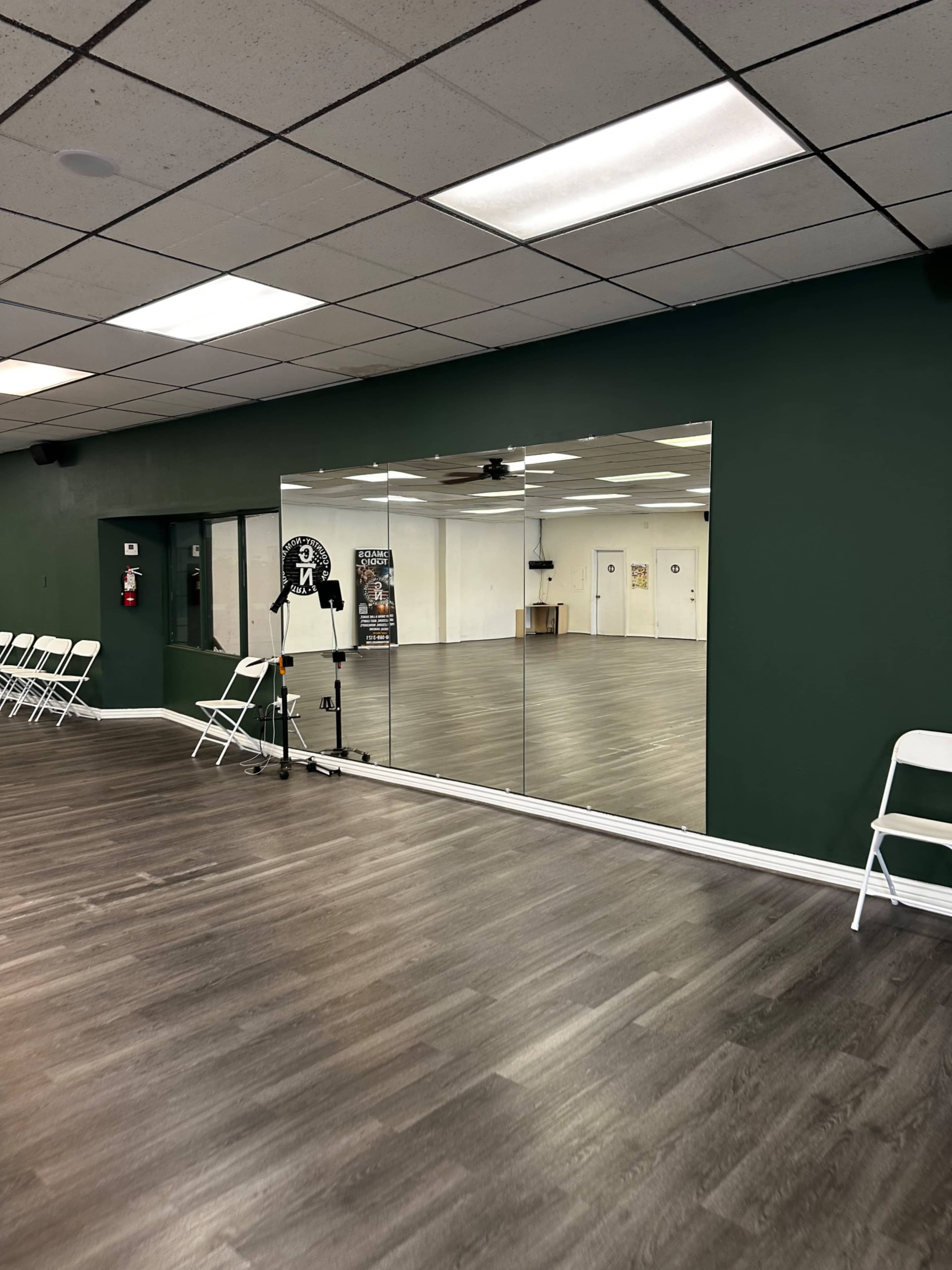 A room with a dark green wall and a large mirror reflecting an empty space with chairs and wooden flooring.