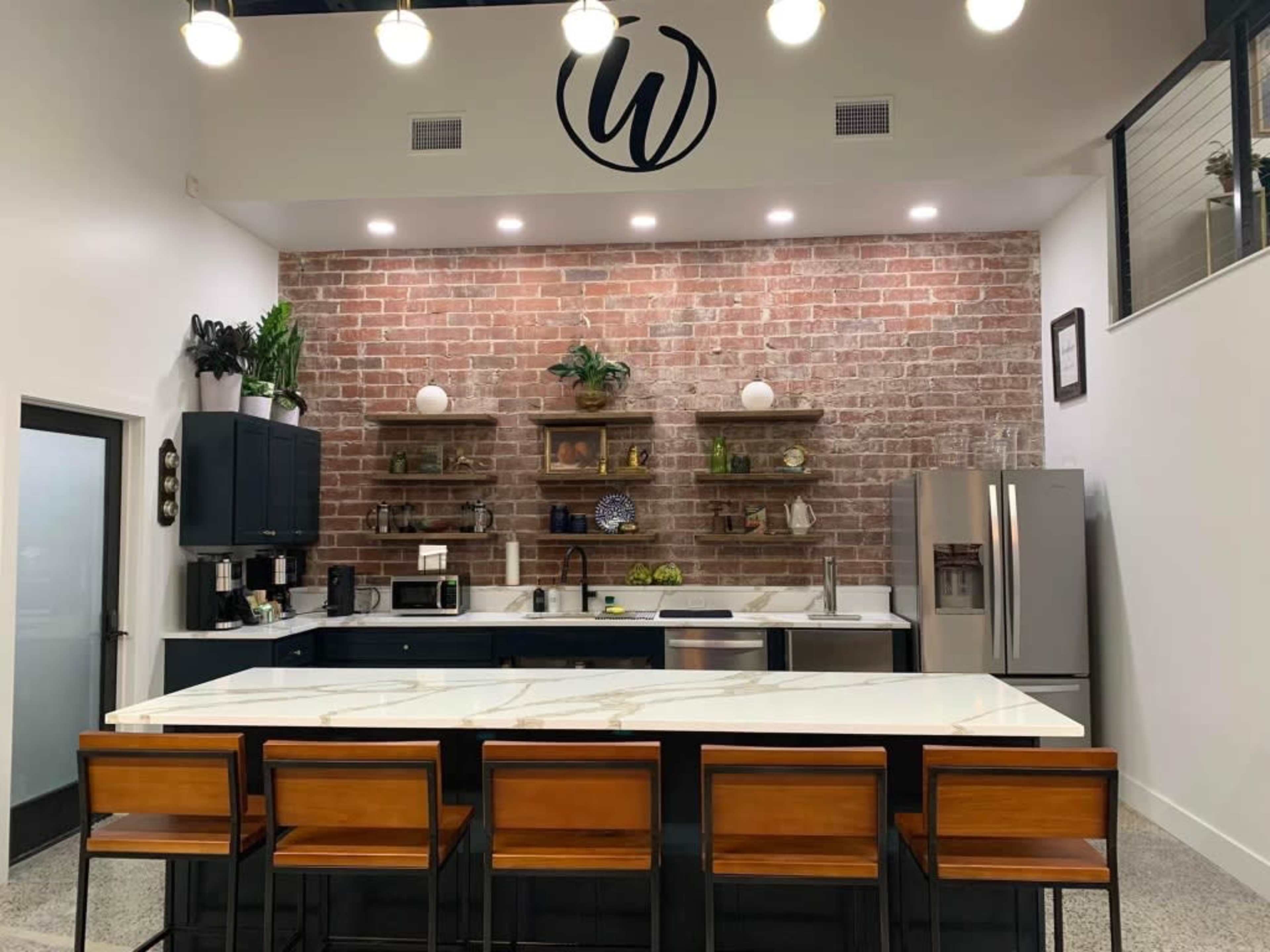 The image features a modern kitchen with a brick accent wall, dark cabinetry, open shelves, and a large island with wooden bar stools.