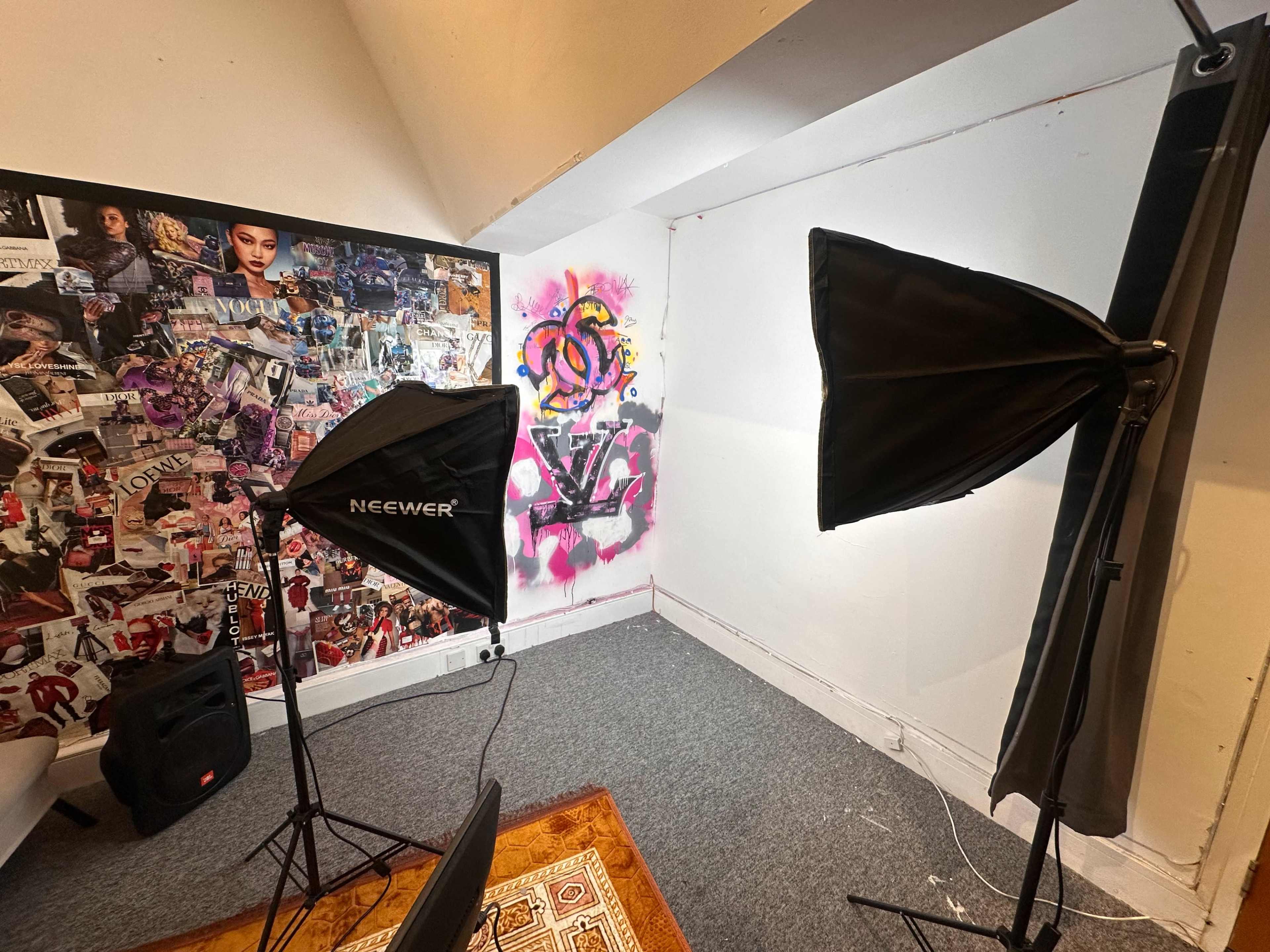 The image shows a small studio or room featuring a collage wall of images, two softbox lights positioned on either side, and a colorful graffiti piece on one wall.