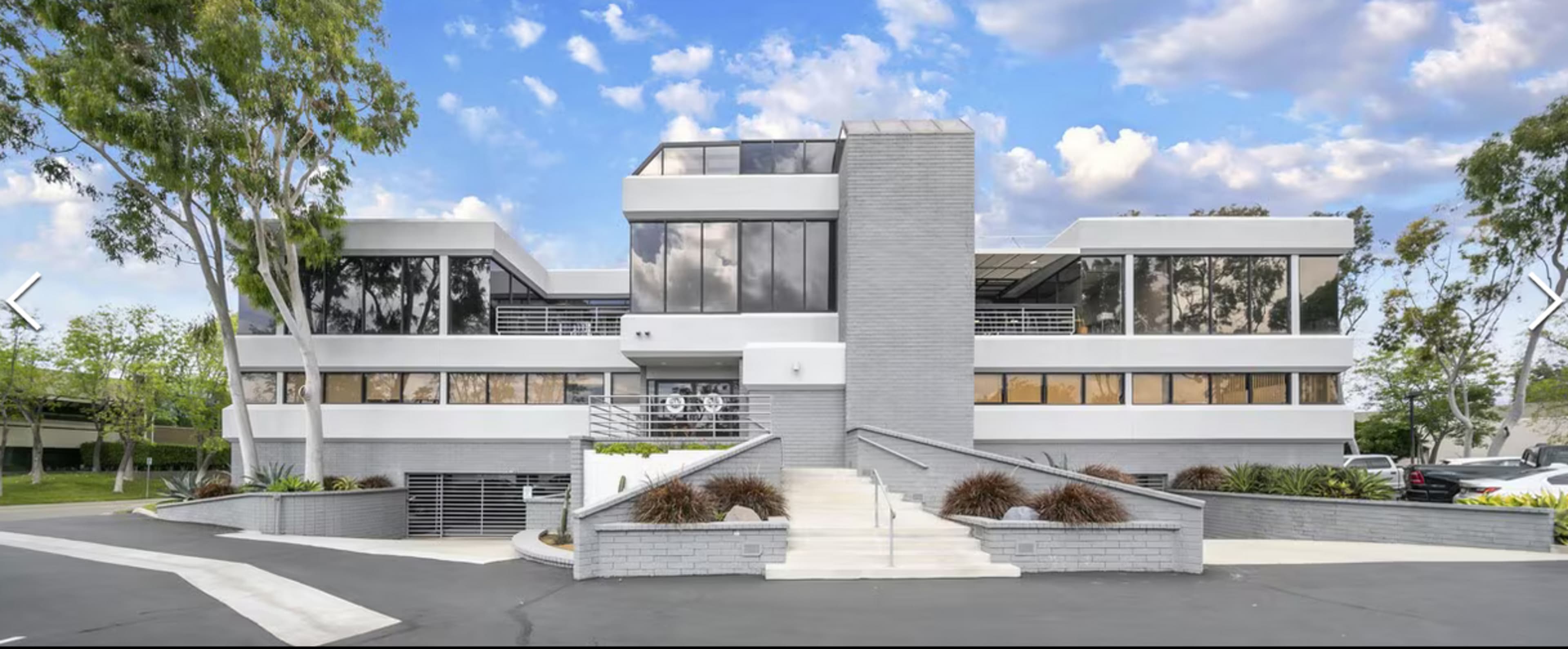 The image shows a modern two-story building with large windows and a minimalist design, surrounded by landscaped greenery and a parking area.