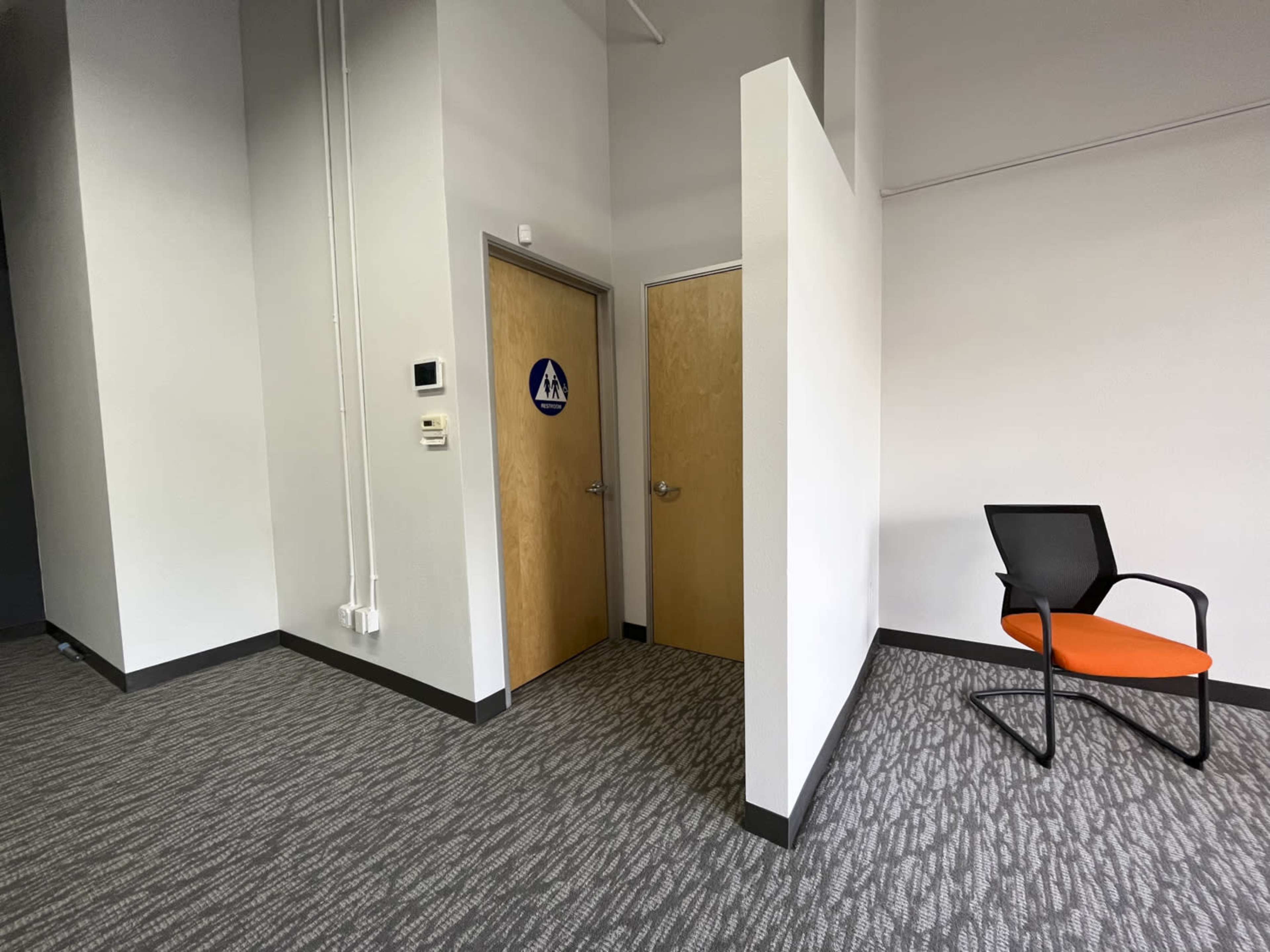 A corner of an indoor space featuring two restroom doors and an orange chair against a gray patterned carpet.