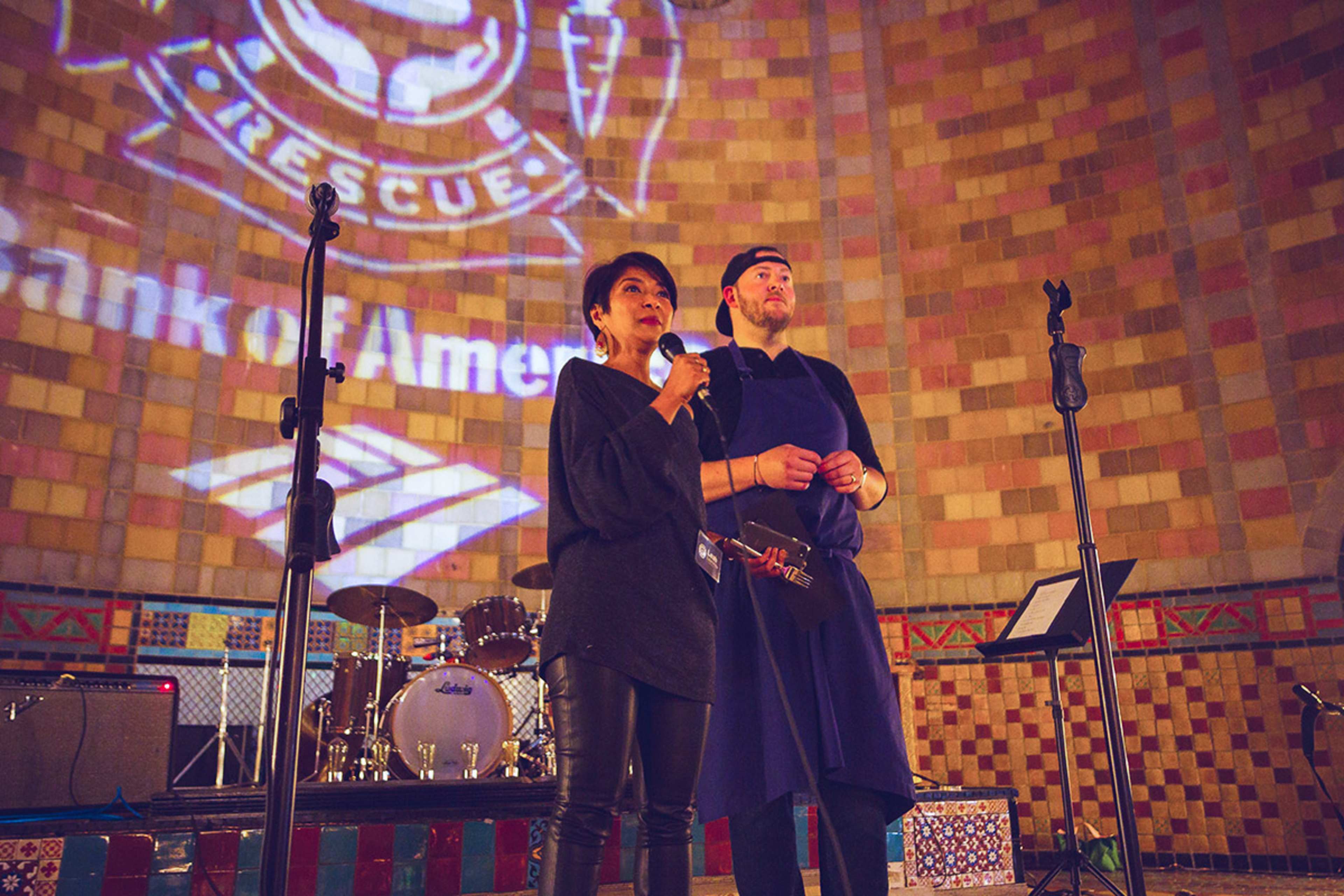 A man and a woman stand on a stage with microphones in front of a projected logo, while musical instruments and a decorated backdrop are visible behind them.