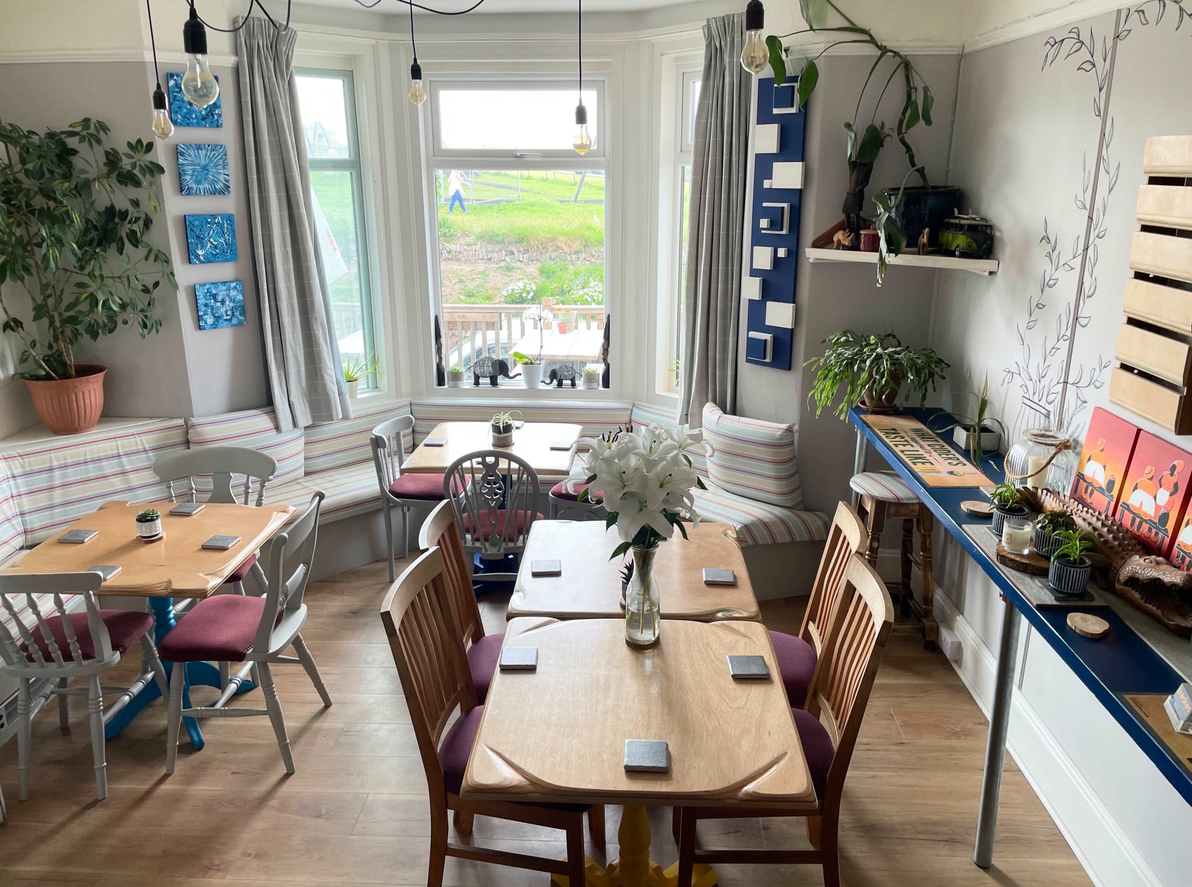 A cozy dining area with wooden tables and chairs, surrounded by large windows and decorated with potted plants and wall art.