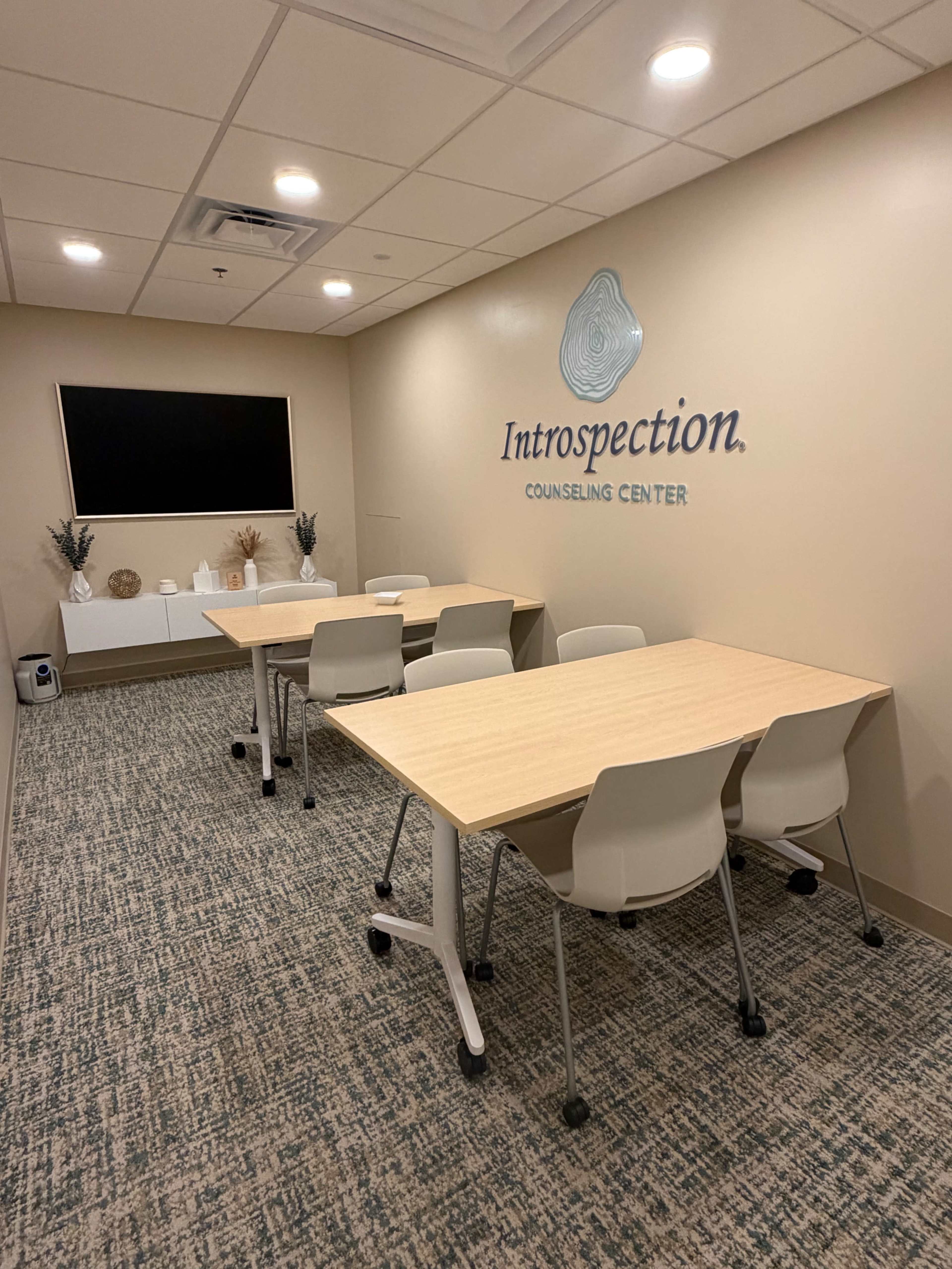 The image shows a counseling center meeting room with several light wooden tables and white chairs, a large wall-mounted screen, and a calming beige color scheme.