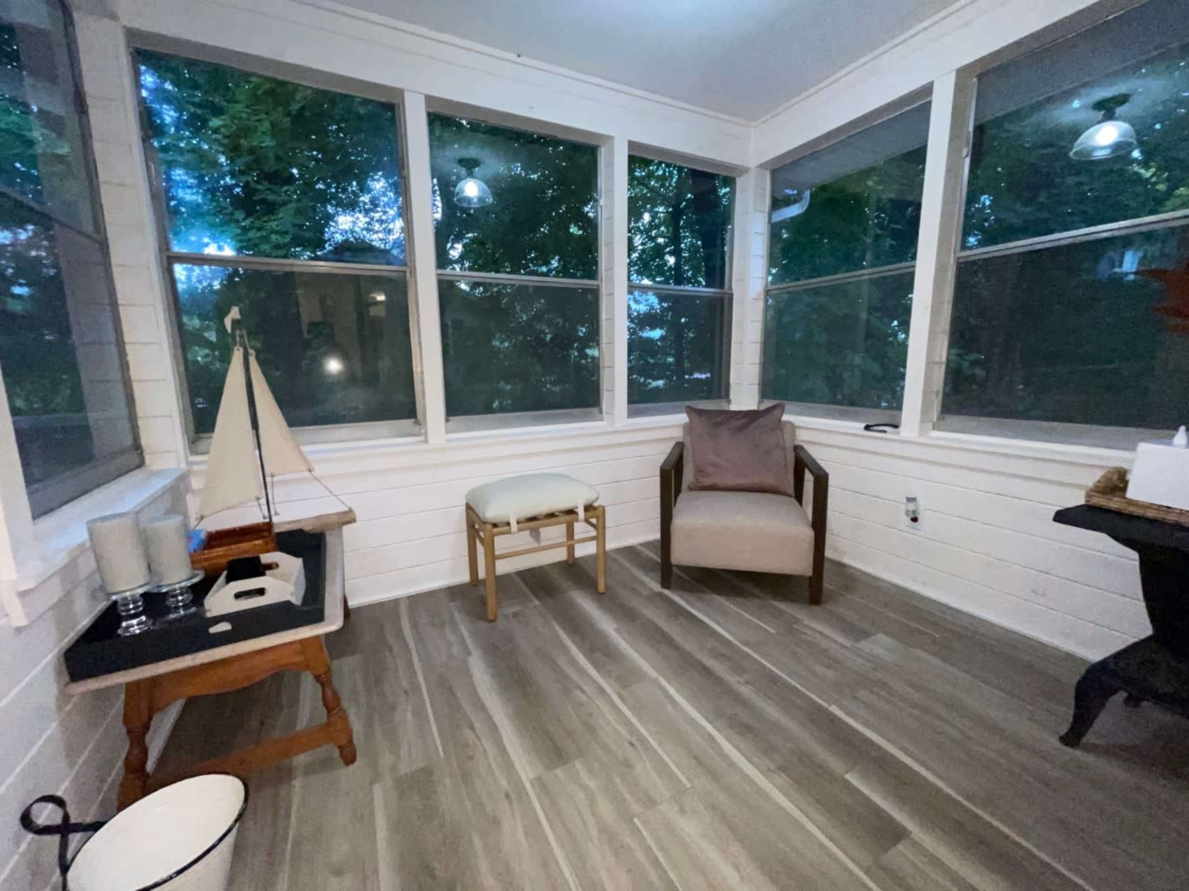 A bright, enclosed porch featuring large windows, a wooden sailboat on a table, a light-colored chair, and a dark side table.