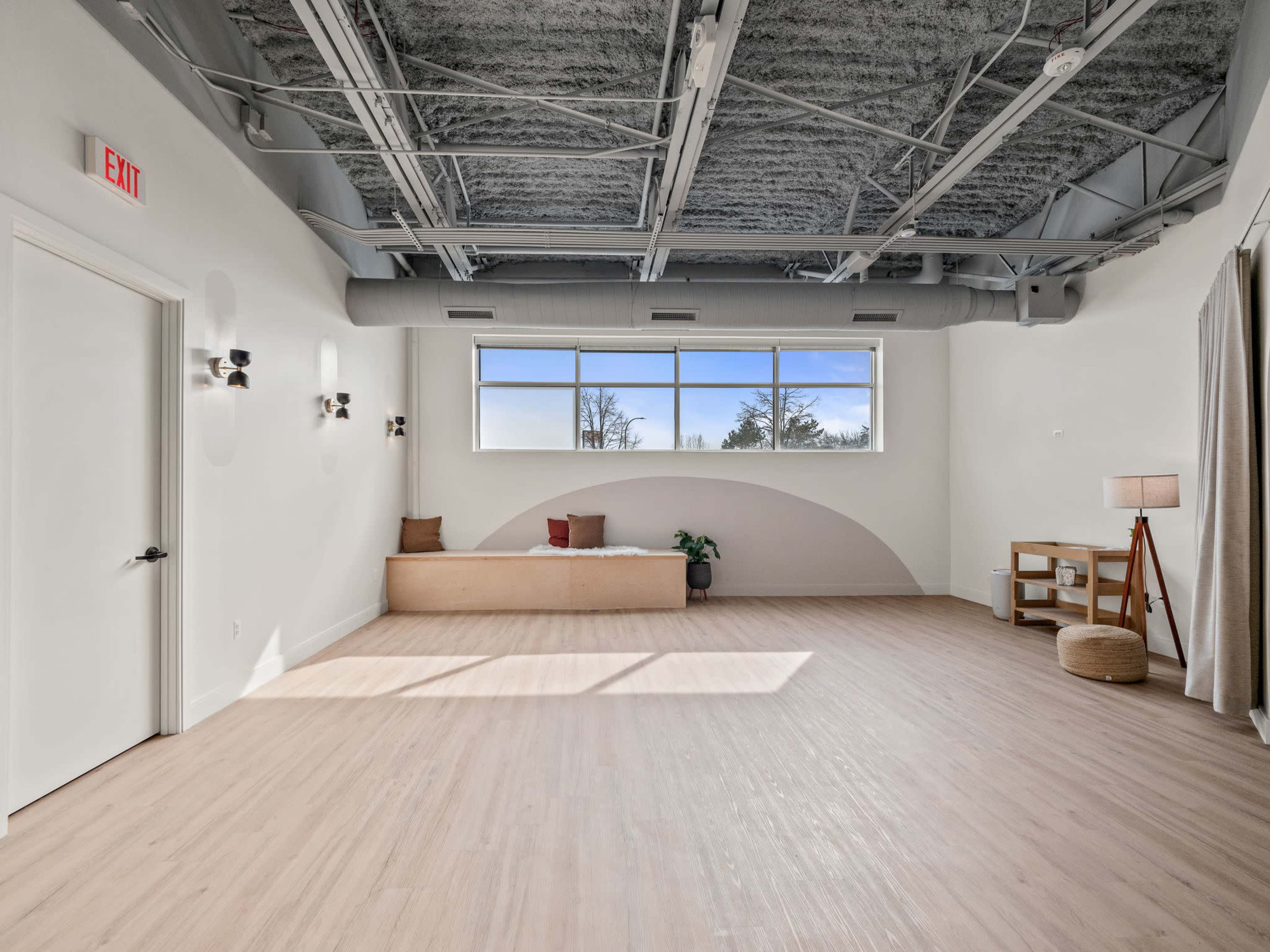 A spacious, well-lit room features a large window, wooden bench, and minimal furniture against a light-colored wall.