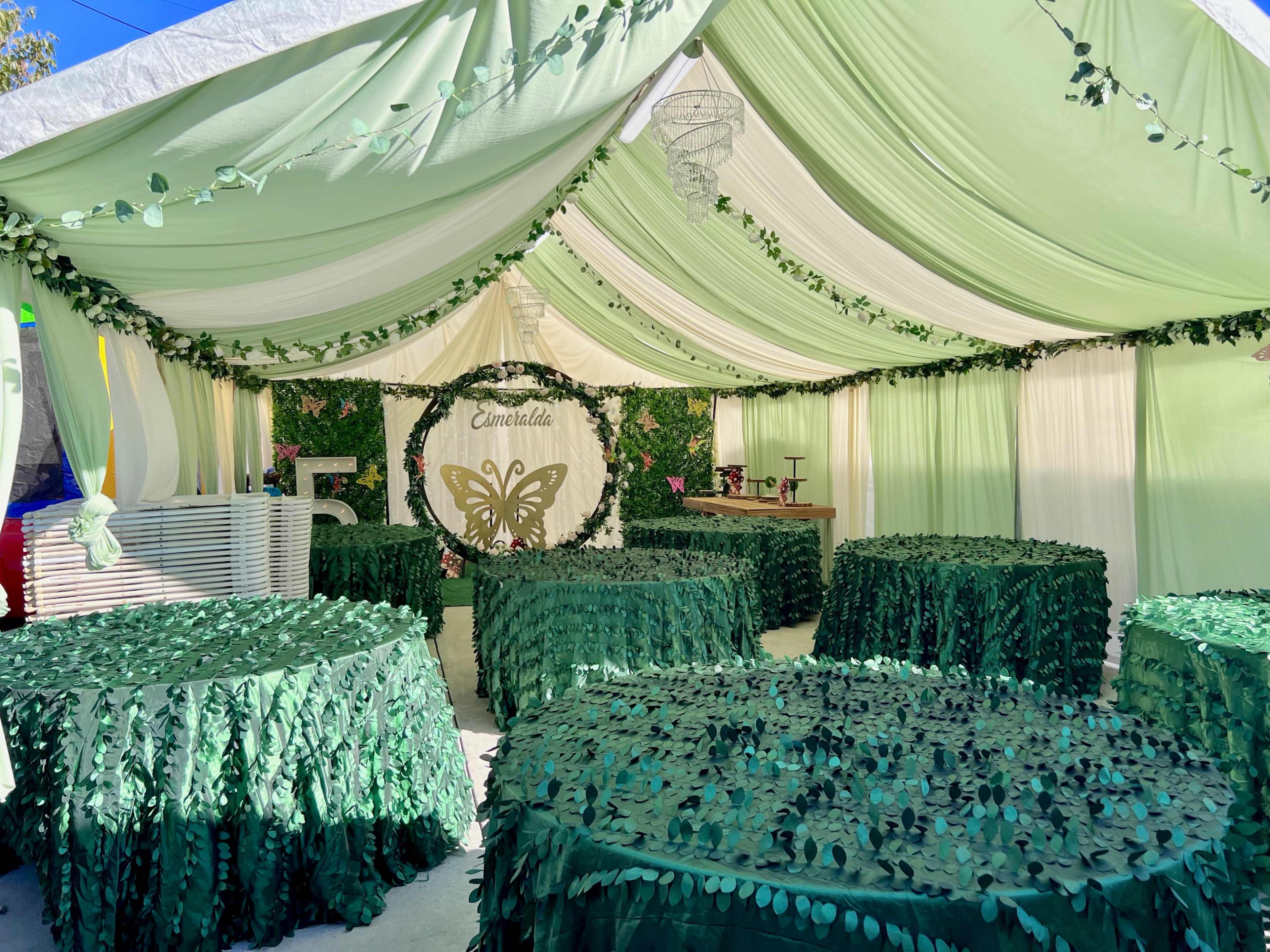 A decorated indoor space with green fabric drapery, leaf-themed table covers, and a butterfly emblem on the backdrop.