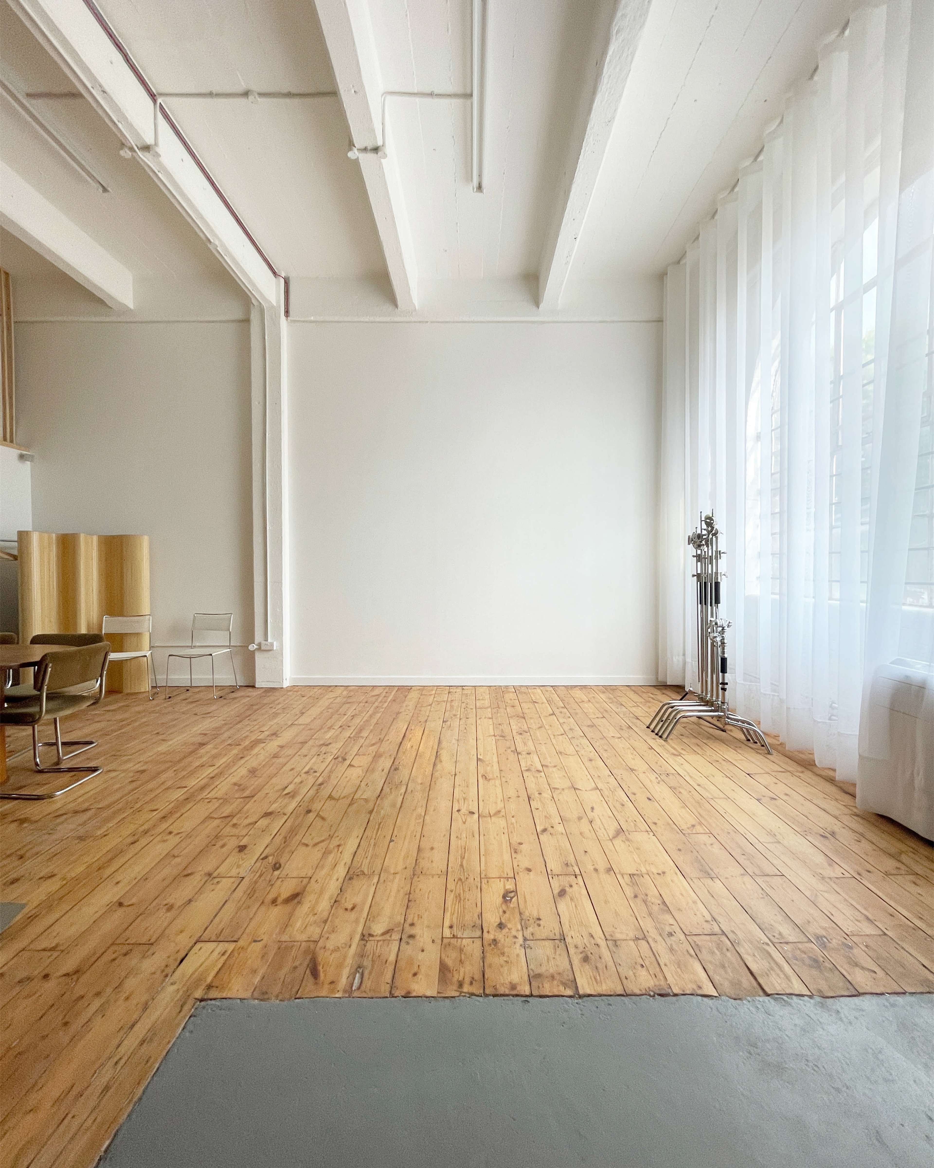 A spacious, empty room features wooden flooring, large windows with sheer curtains, and minimal furniture.