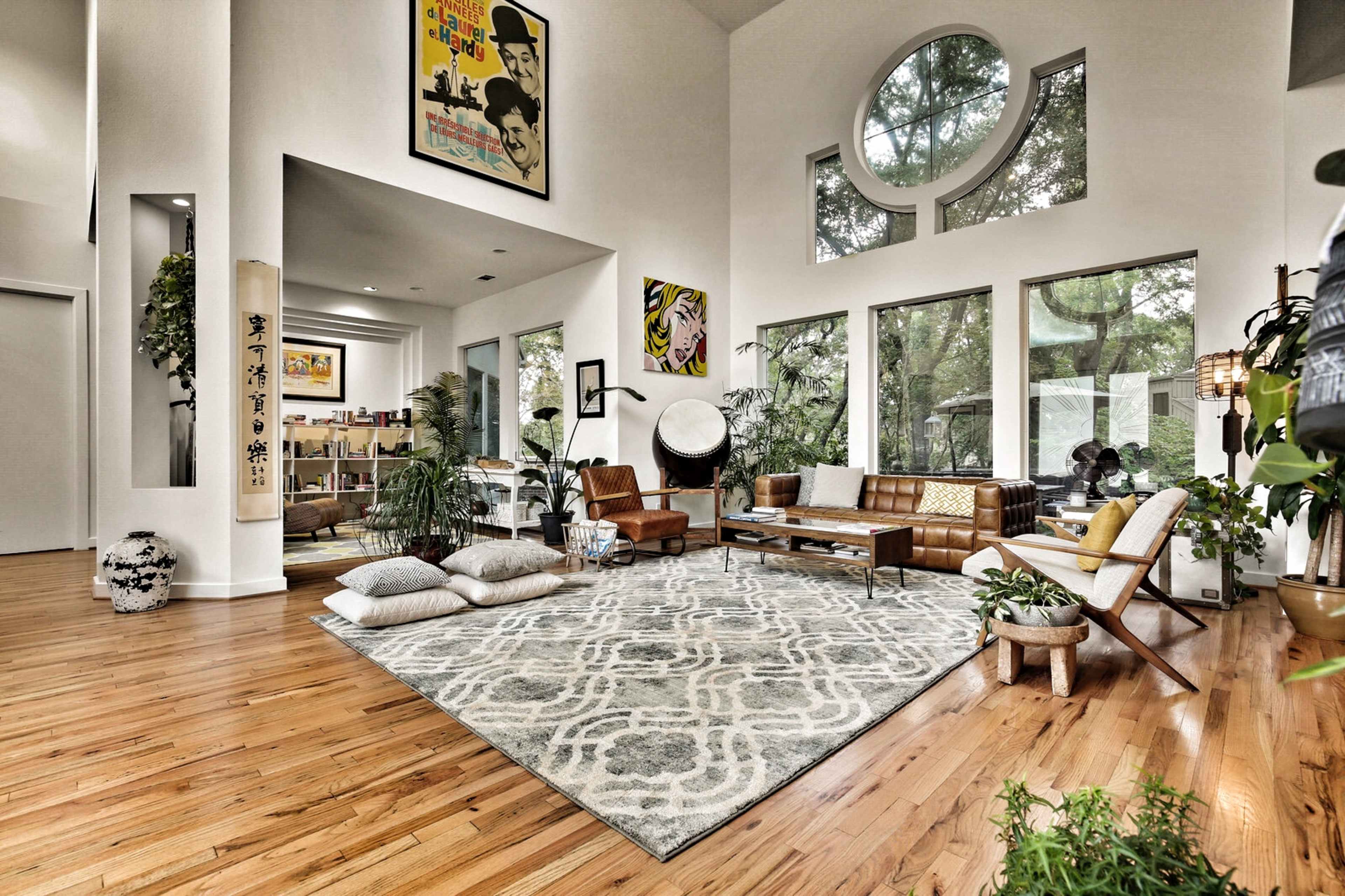 A spacious living area featuring large windows, a patterned rug, wooden flooring, plants, modern furniture, and vibrant artwork on the walls.
