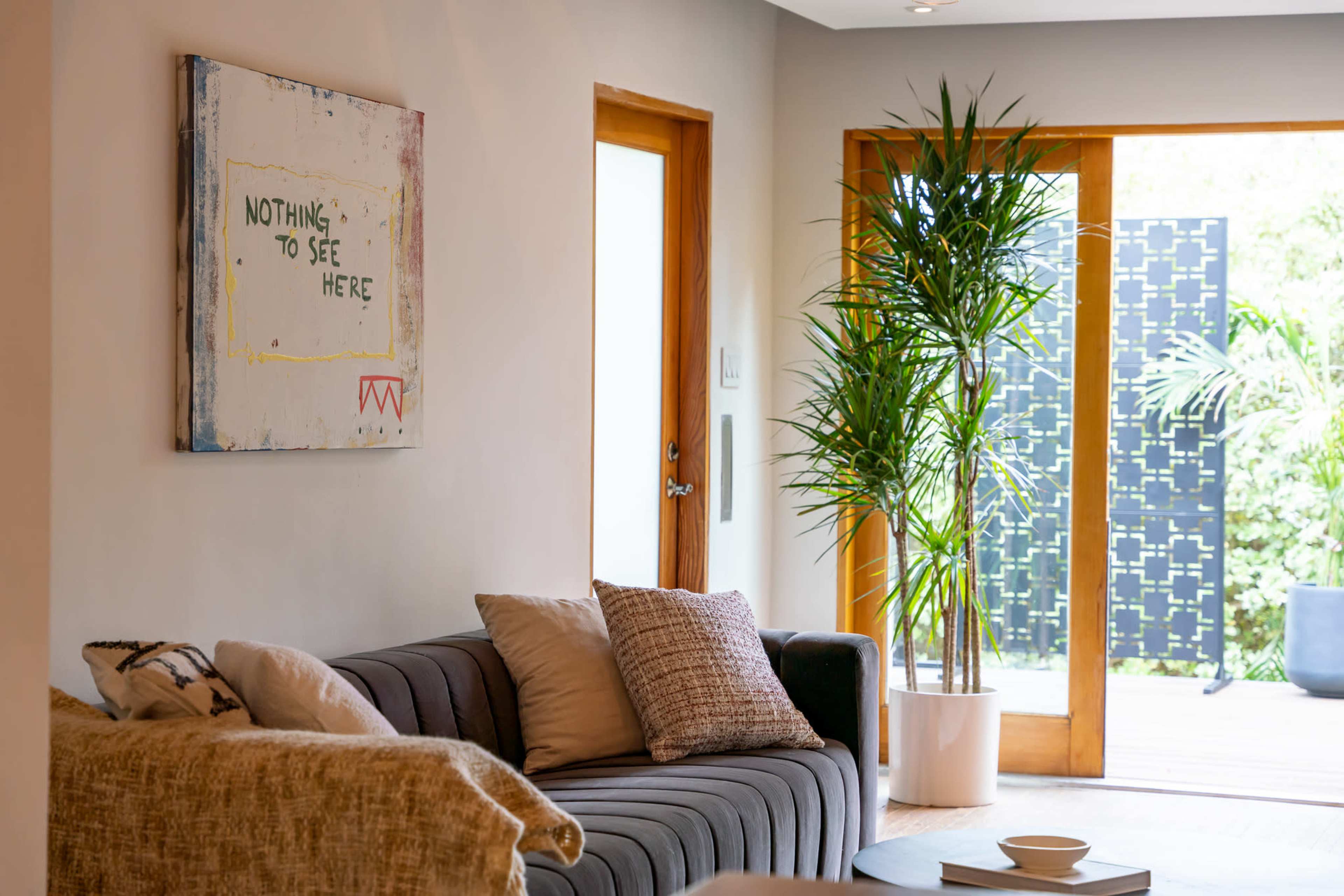 A living room features a gray sofa with decorative pillows, a palm plant in a white pot, and a painting on the wall that reads "NOTHING TO SEE HERE."