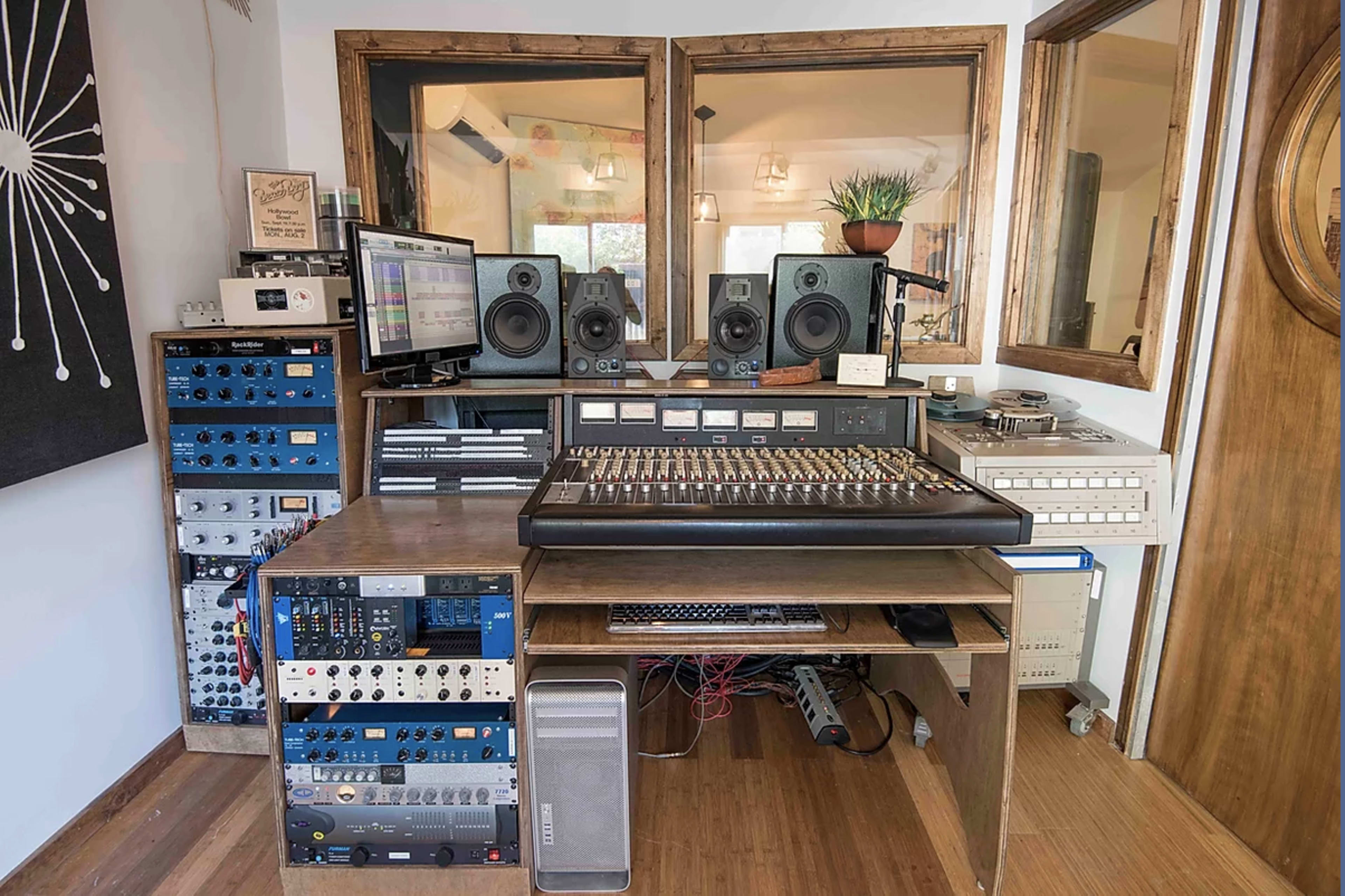 The image shows a recording studio setup featuring a large mixing console, audio equipment racks, speakers, a computer monitor, and a wood-paneled interior.