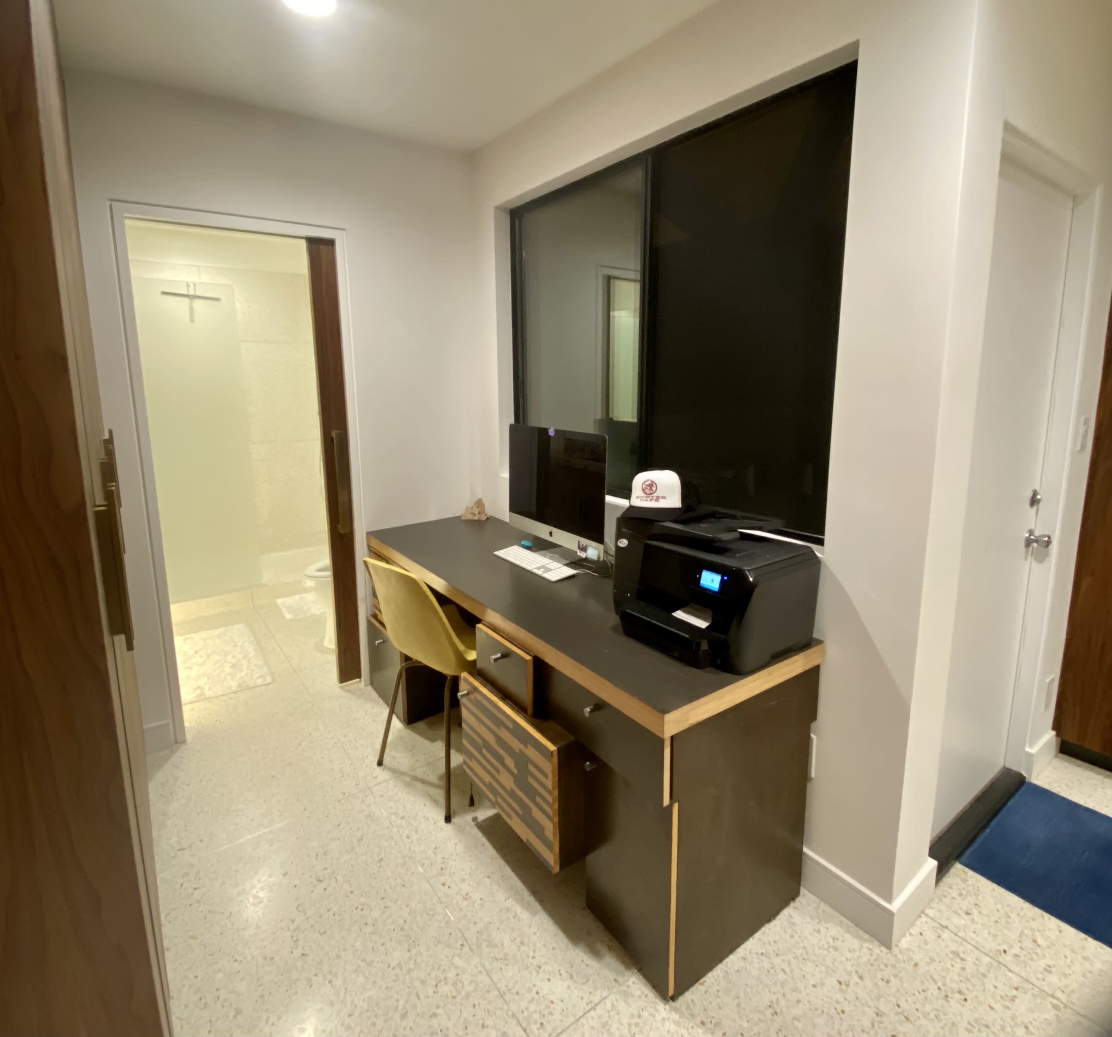 The image shows a small workspace with a desk, computer, printer, and a bathroom partially visible in the background.