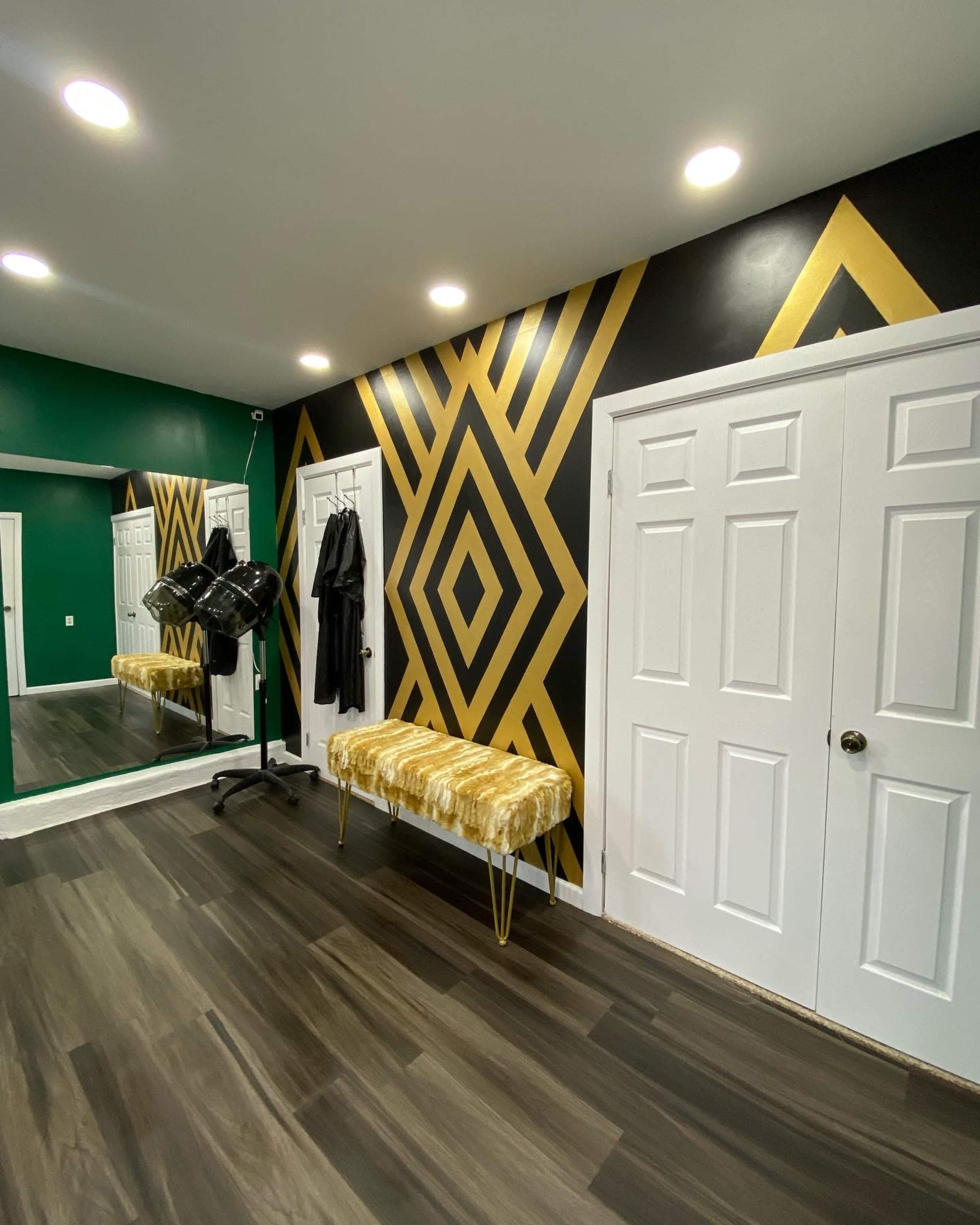 The room features a black and gold geometric patterned wall, a large mirror, a plush gold bench, and two closed white doors.