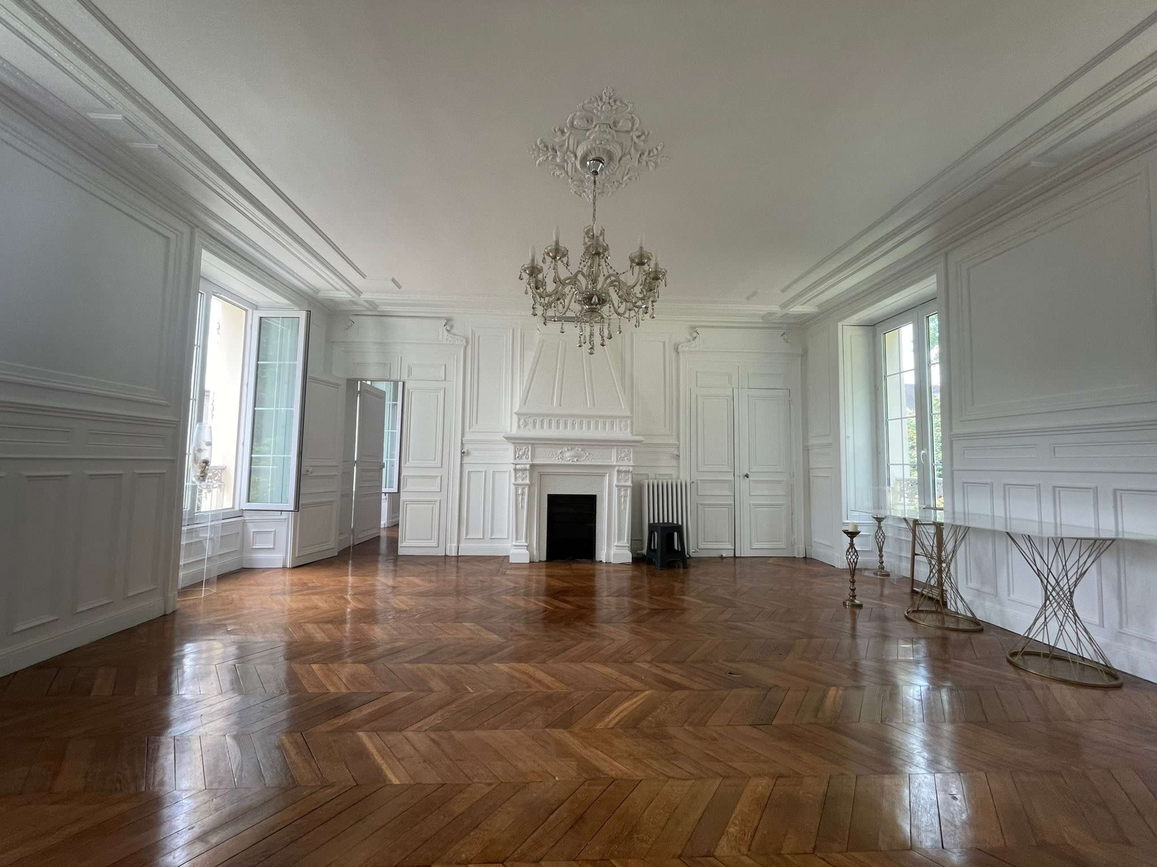 A spacious, empty room with white walls, a decorative chandelier, and a polished wooden floor arranged in a herringbone pattern.