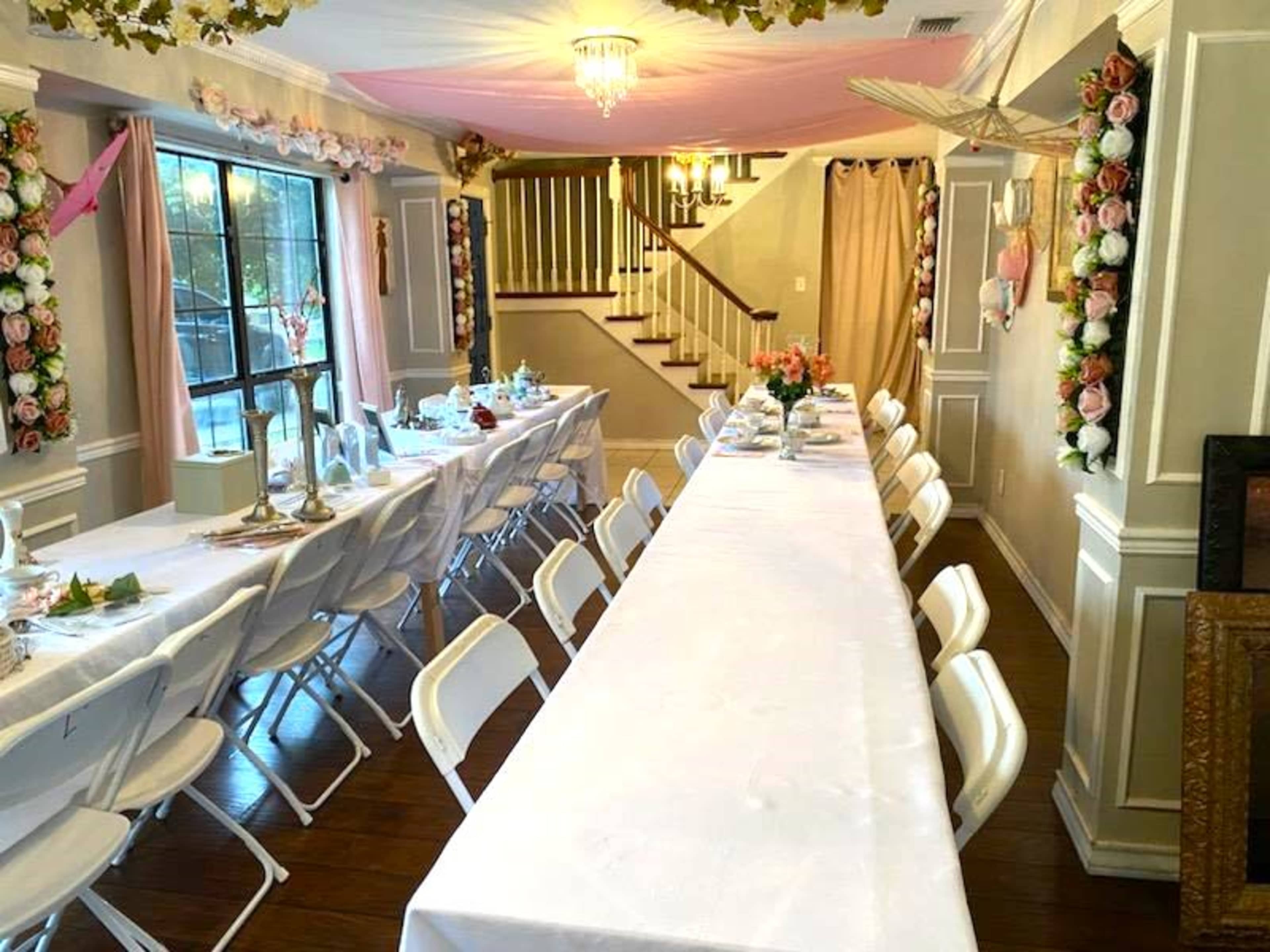 A dining area features two long tables covered with white tablecloths, surrounded by white chairs, with floral decorations on the walls and a chandelier overhead.