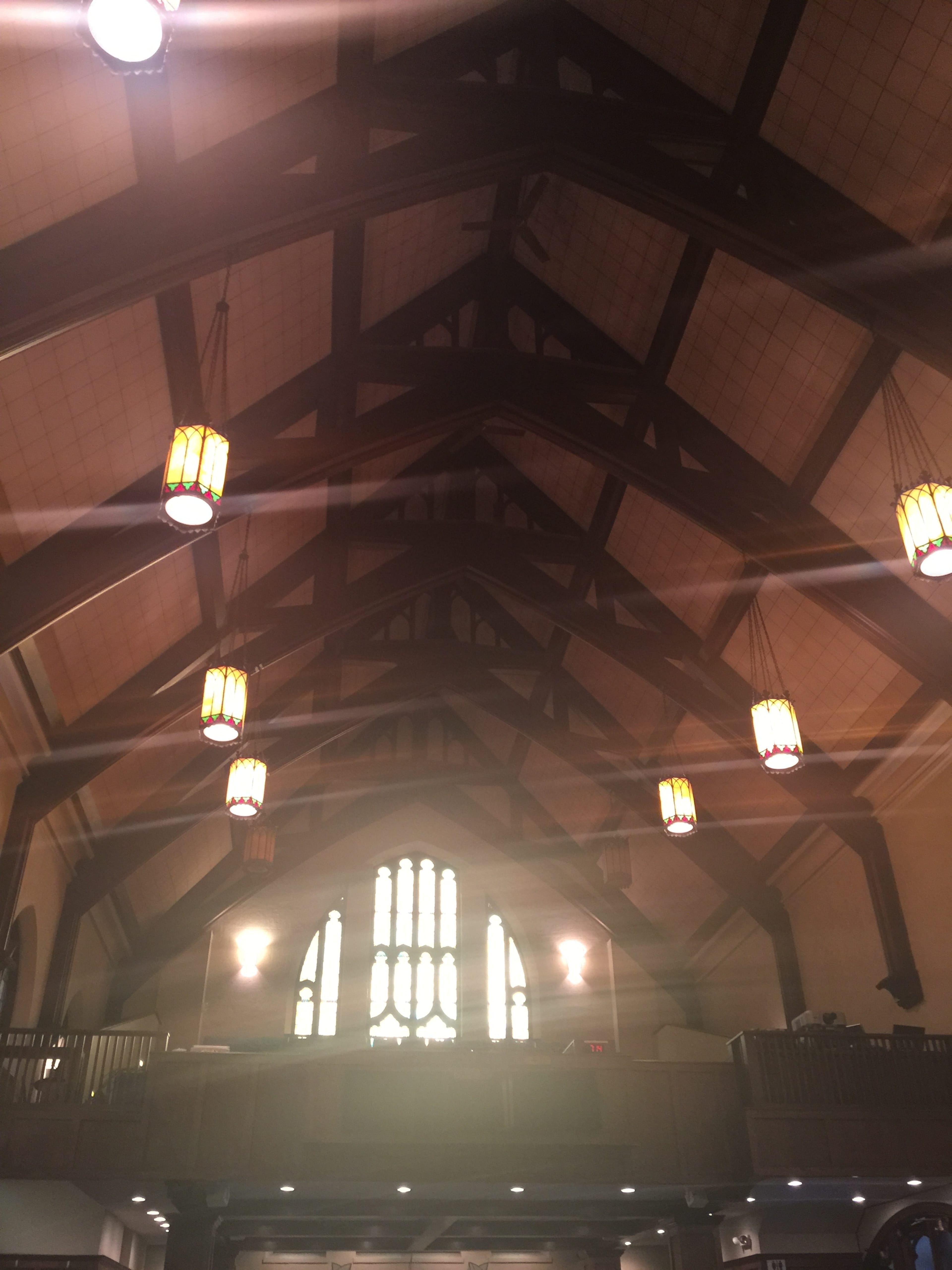 The interior of a church featuring high wooden beams and pendant lights, with a large stained glass window at the back.