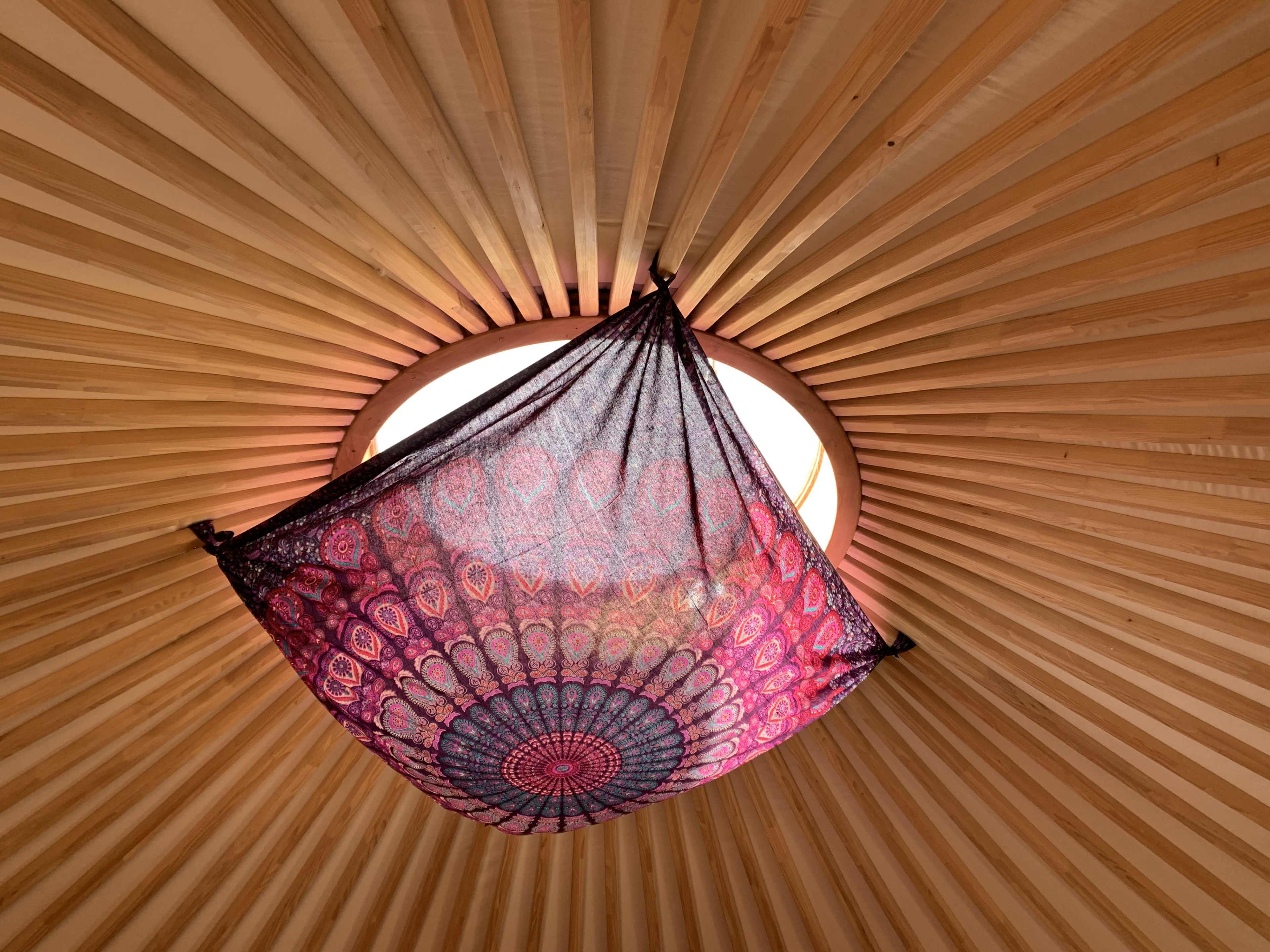 A decorative fabric hangs over a circular skylight in a wooden yurt, surrounded by radiating beams.