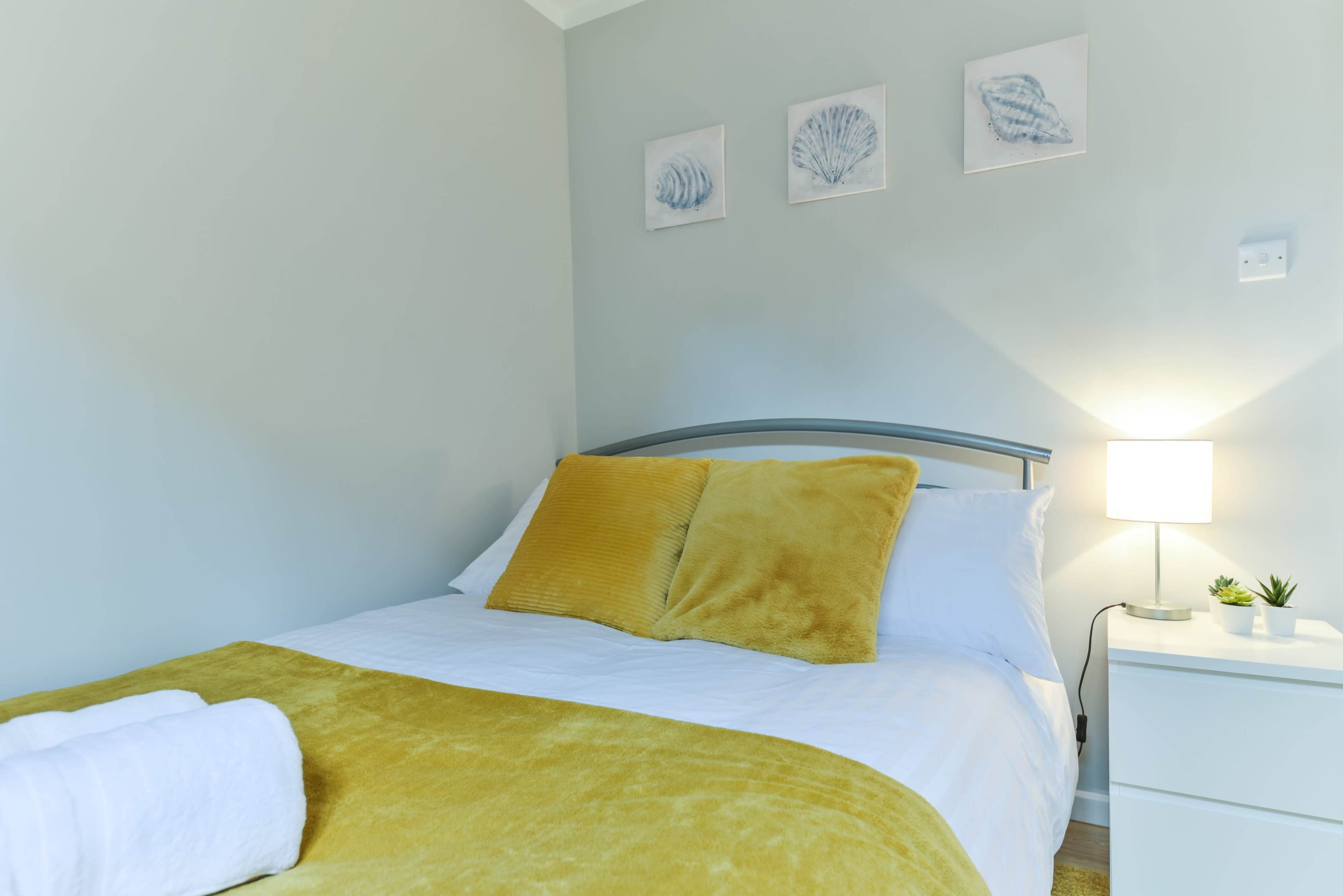 A neatly made bed with yellow cushions is positioned against a light gray wall, accompanied by framed shell artwork and a bedside lamp.