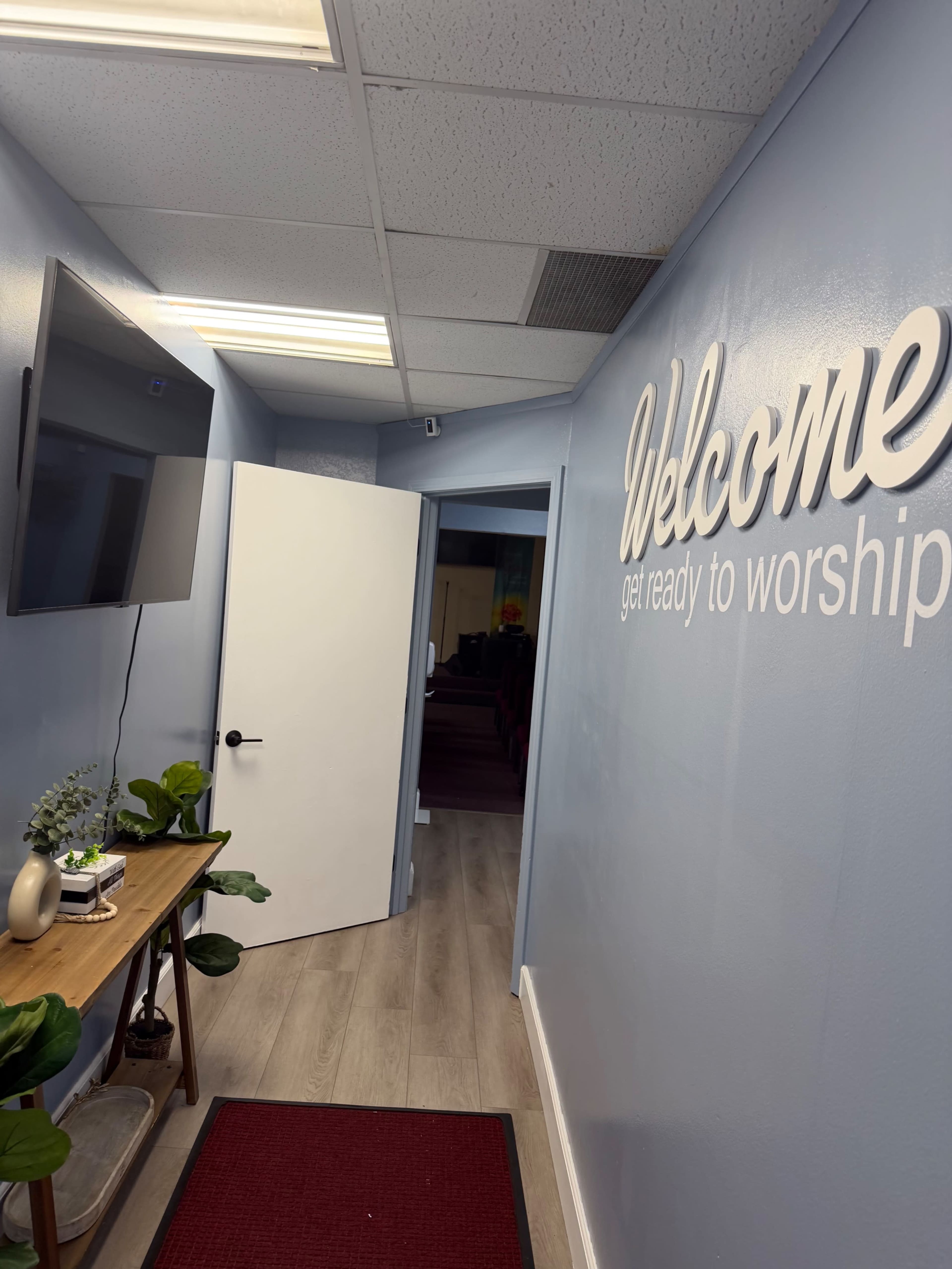 The image shows a hallway leading to a room with a welcoming sign, a television mounted on the wall, and decorative plants on a table.