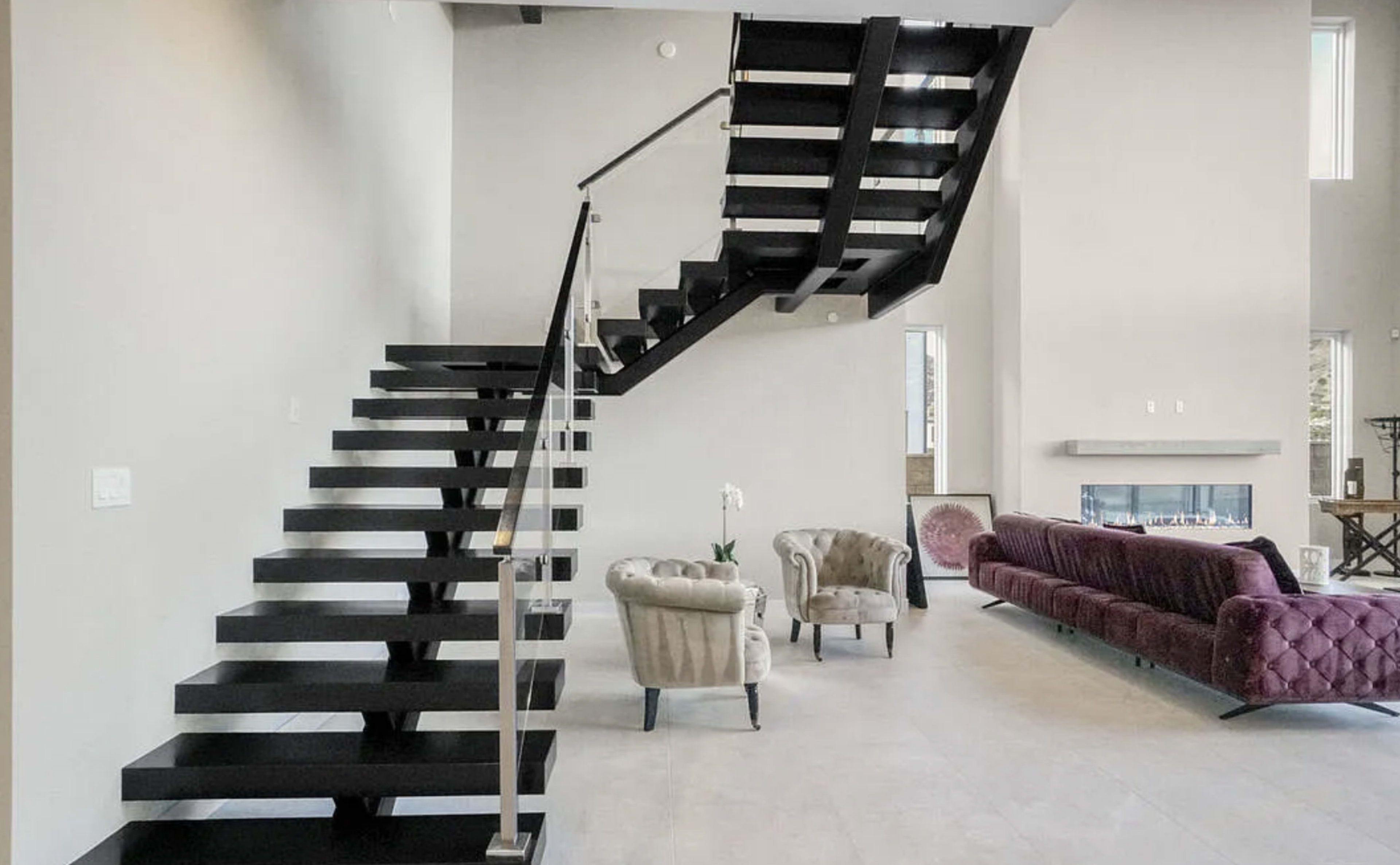 The image shows a modern interior featuring a black, floating staircase, a plush sofa, and two armchairs in a spacious, well-lit living area.