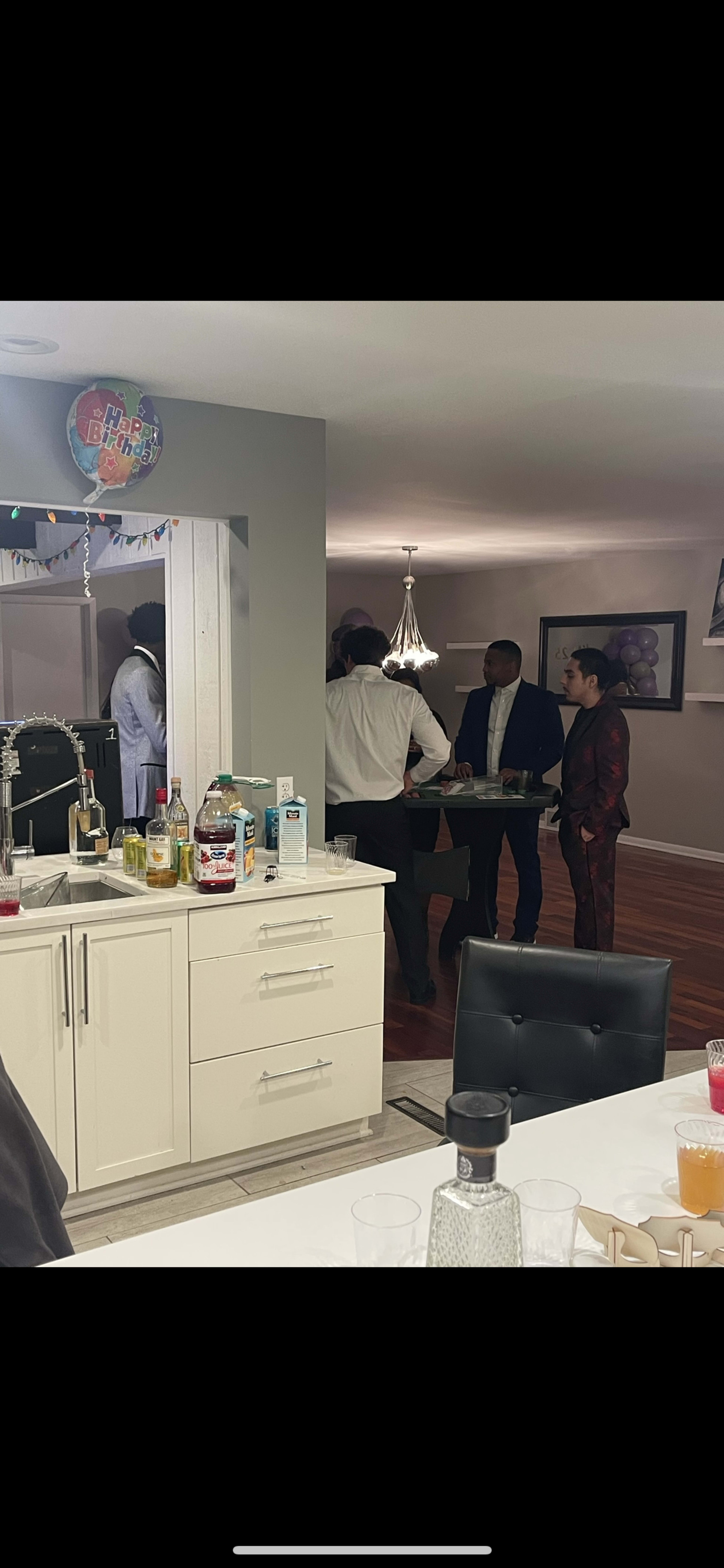 A group of people dressed in formal attire stands around a table in a well-lit kitchen area.